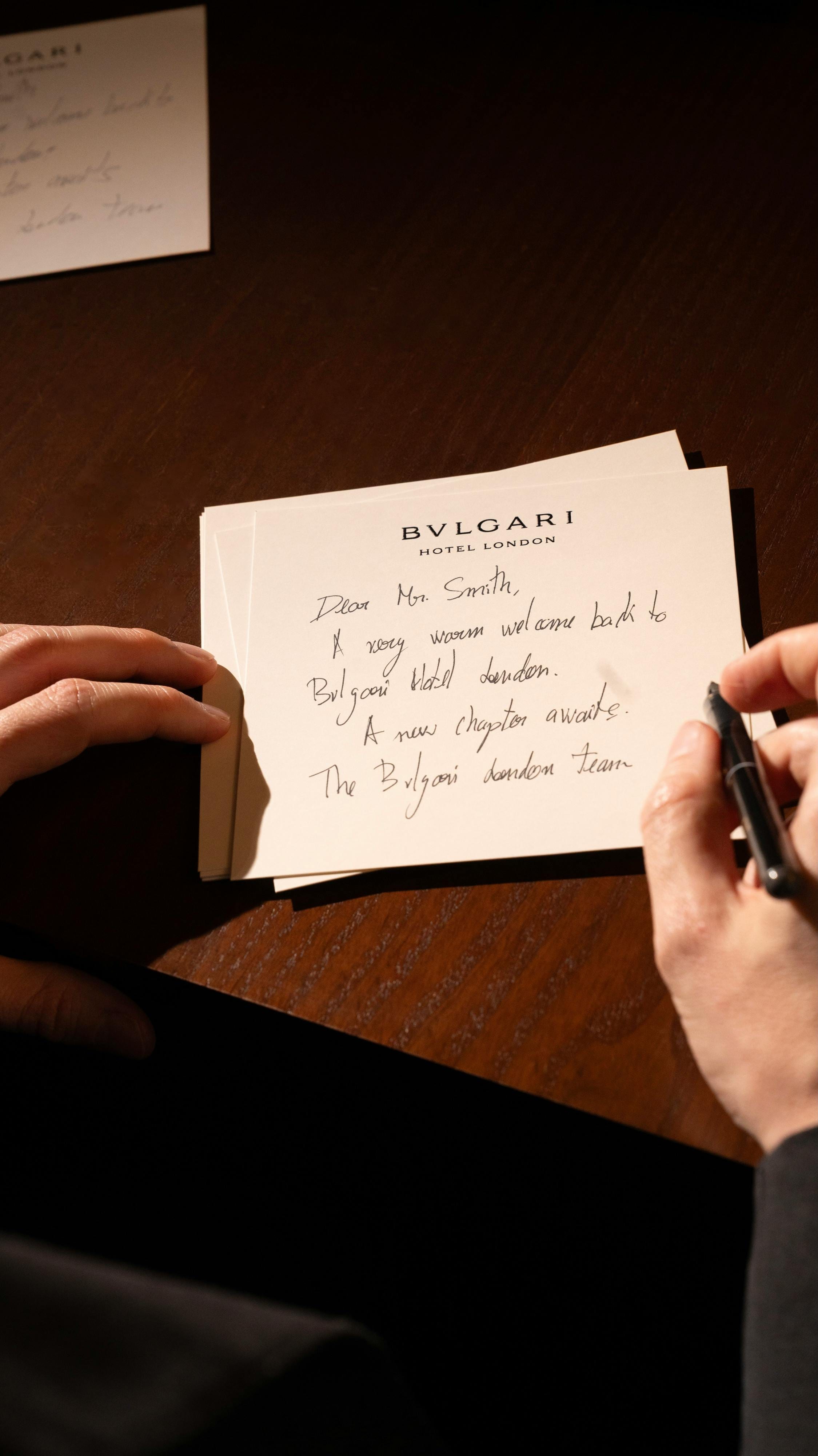Hands writing a welcome note on Bvlgari Hotel London stationery at a dark wood desk