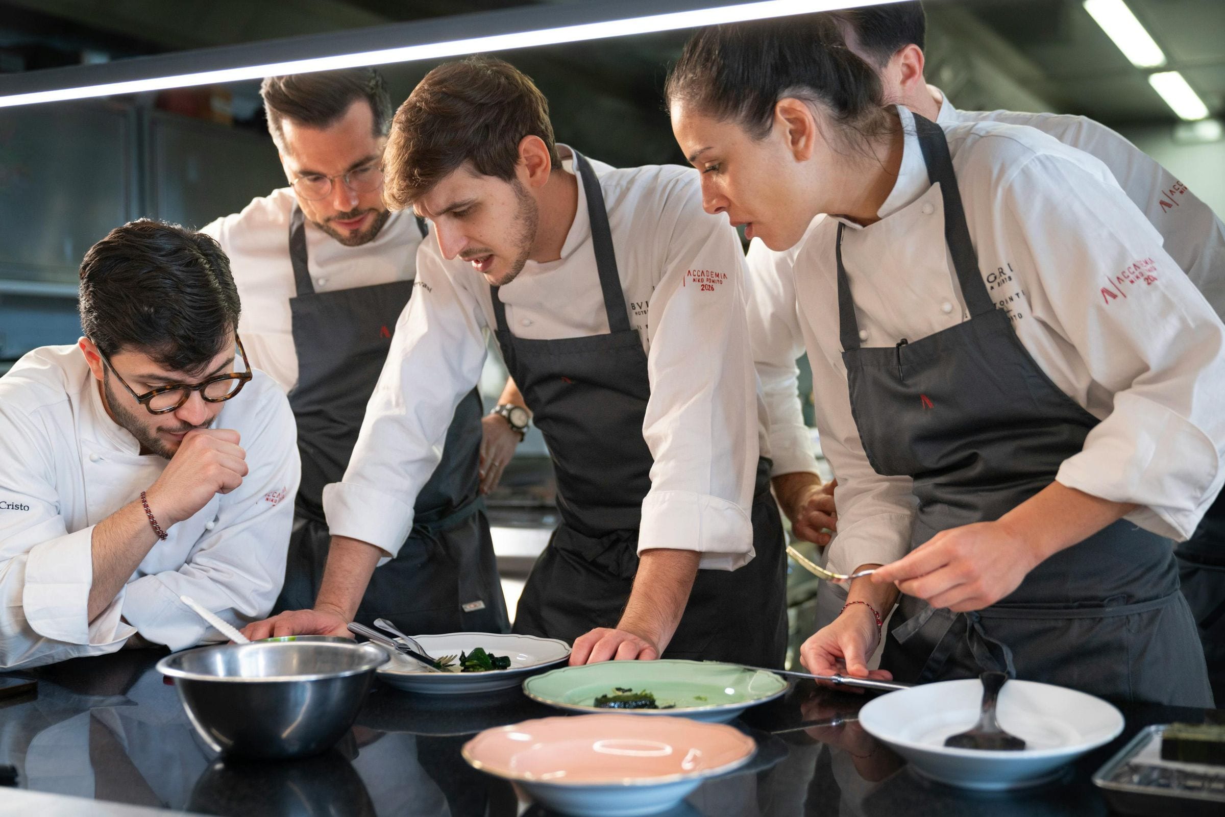 Five chefs in whites leaning over plated dishes at a stainless kitchen pass