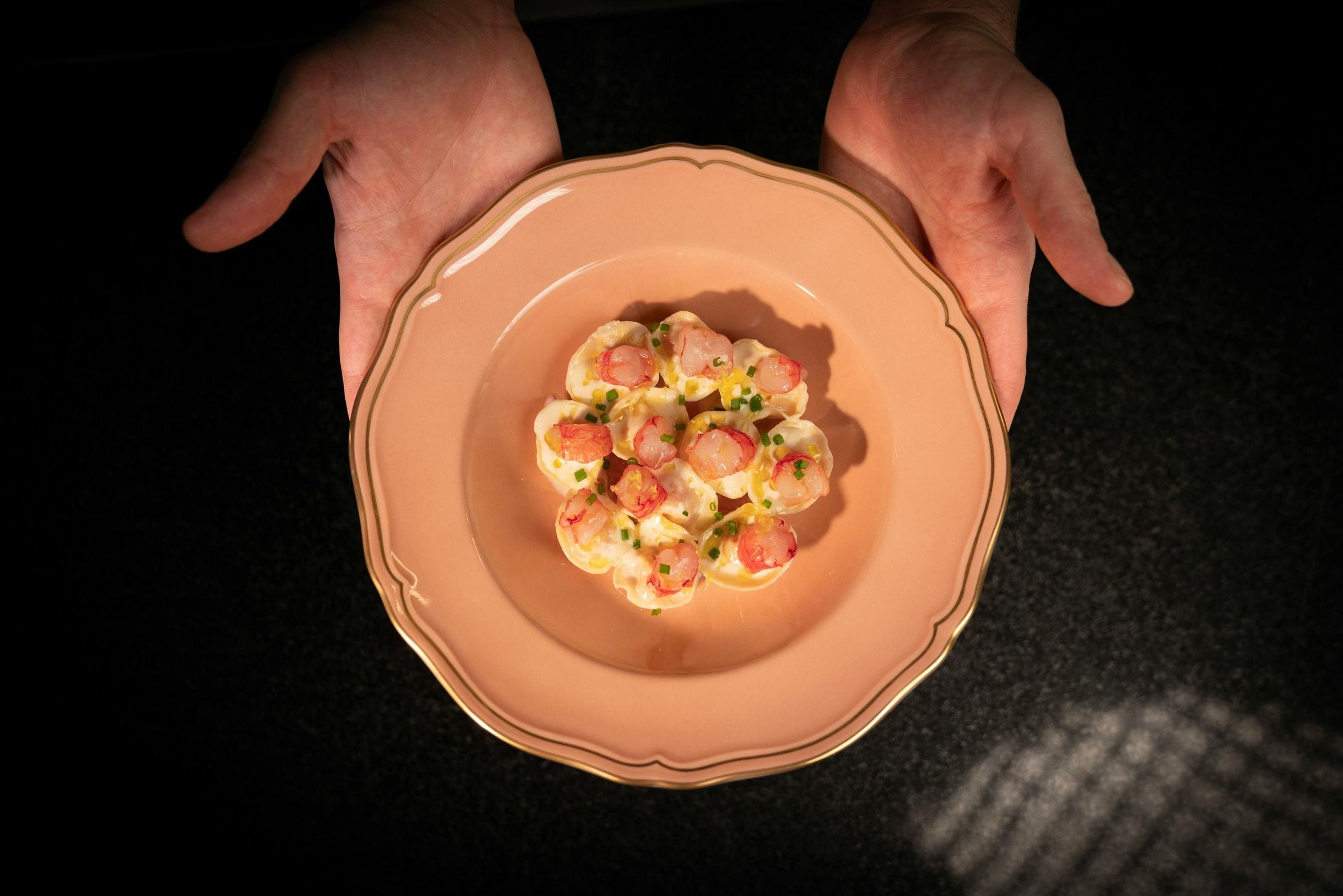 Hands presenting a blush plate of prawn tortelli with chives, seen from above