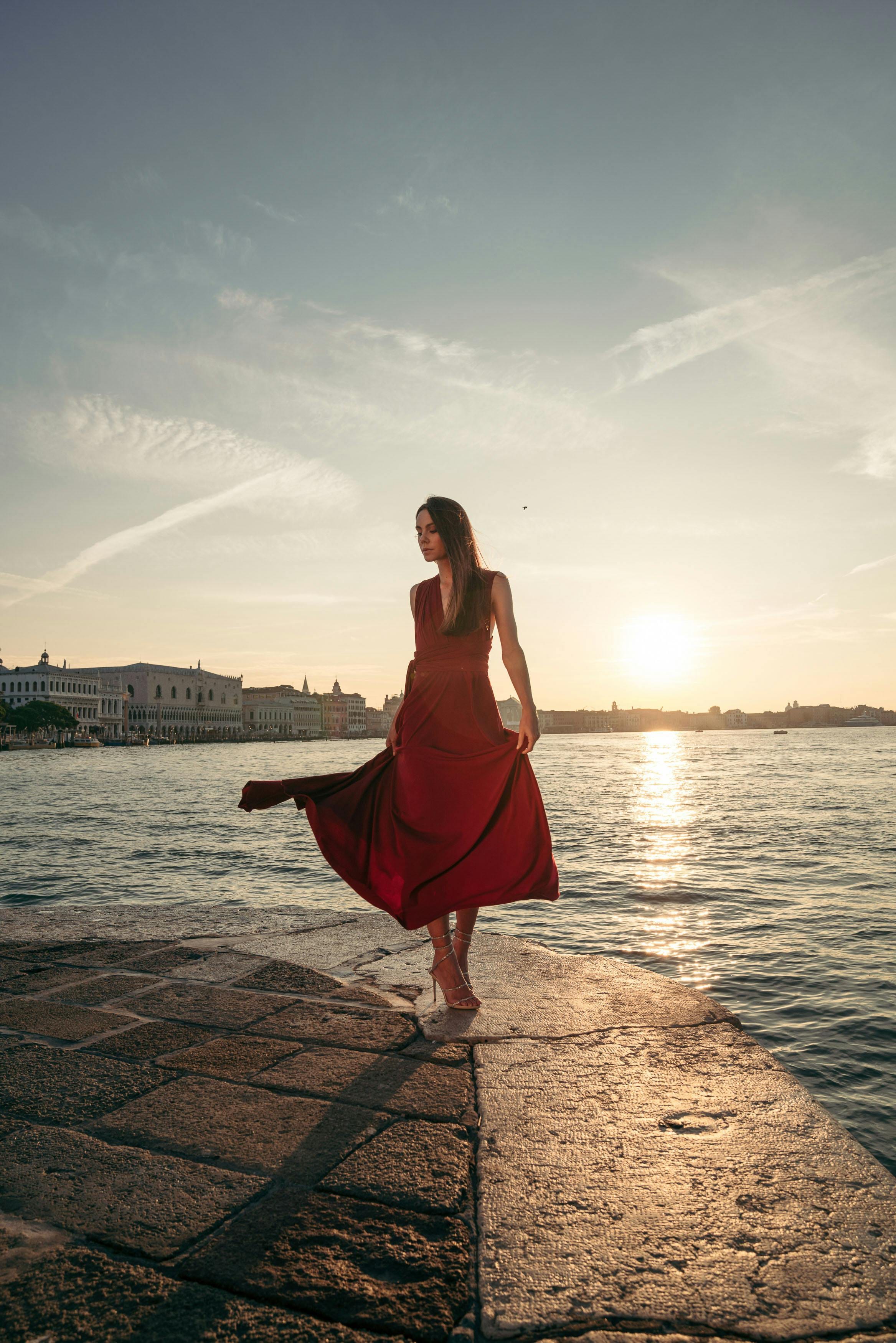 Woman in a red dress twirling on a stone jetty at sunset over the Venice lagoon