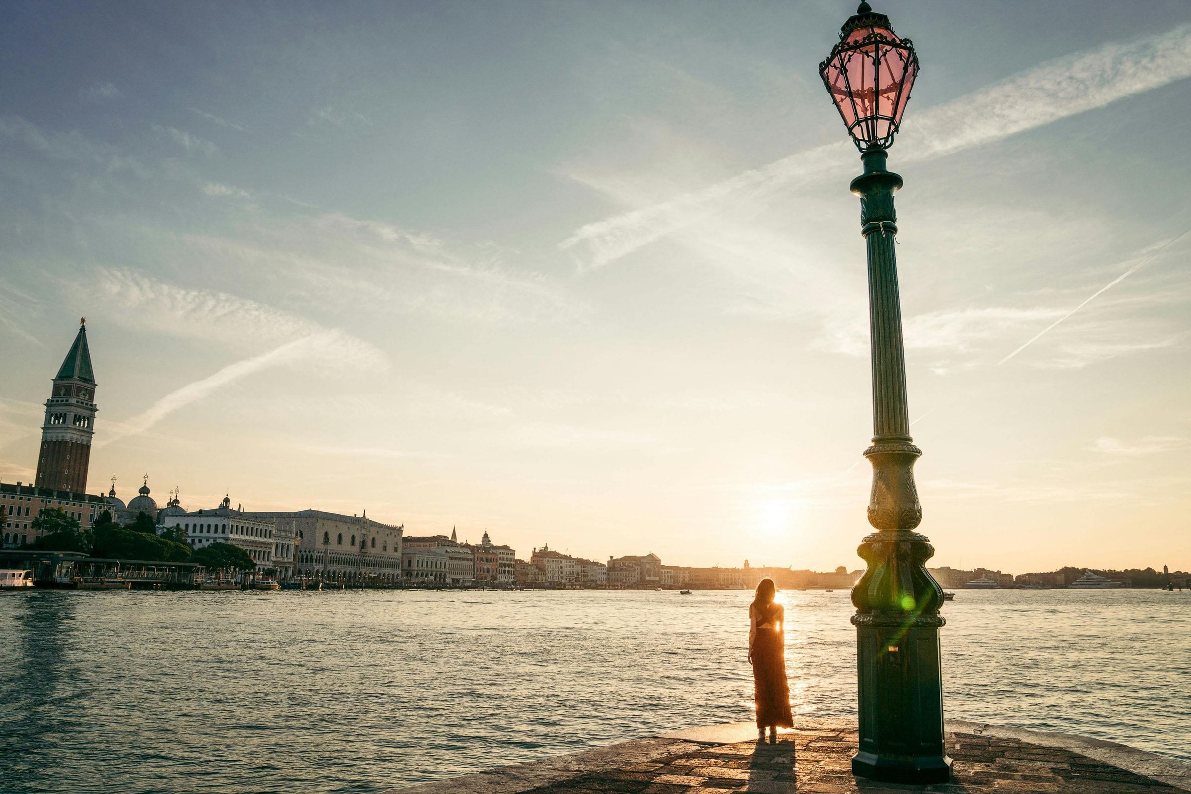 Woman silhouetted by a Venetian lamp post at sunrise, San Marco campanile across the water