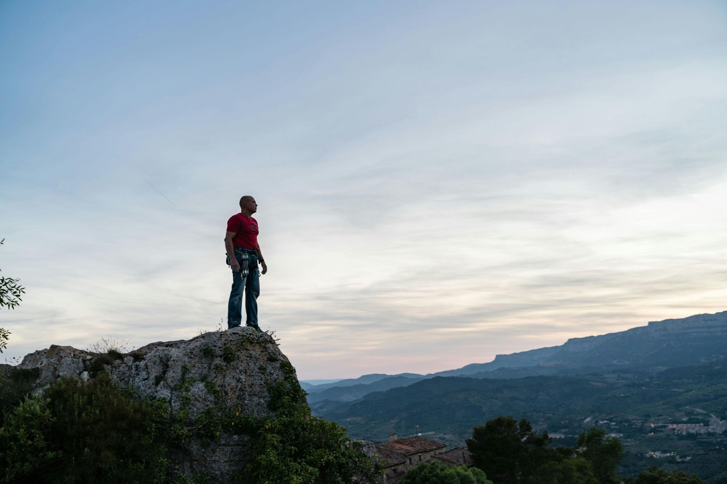 Climber in a red shirt stands on a rocky outcrop above Catalan hills at dusk