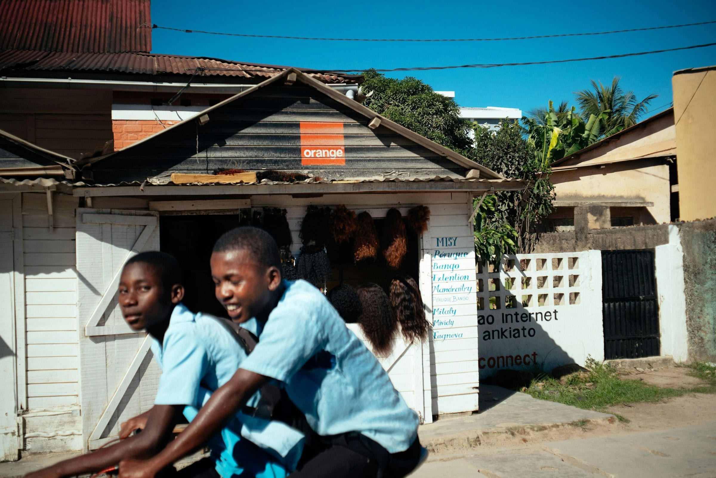 Two schoolboys in blue uniforms cycling past a hair shop with Orange sign