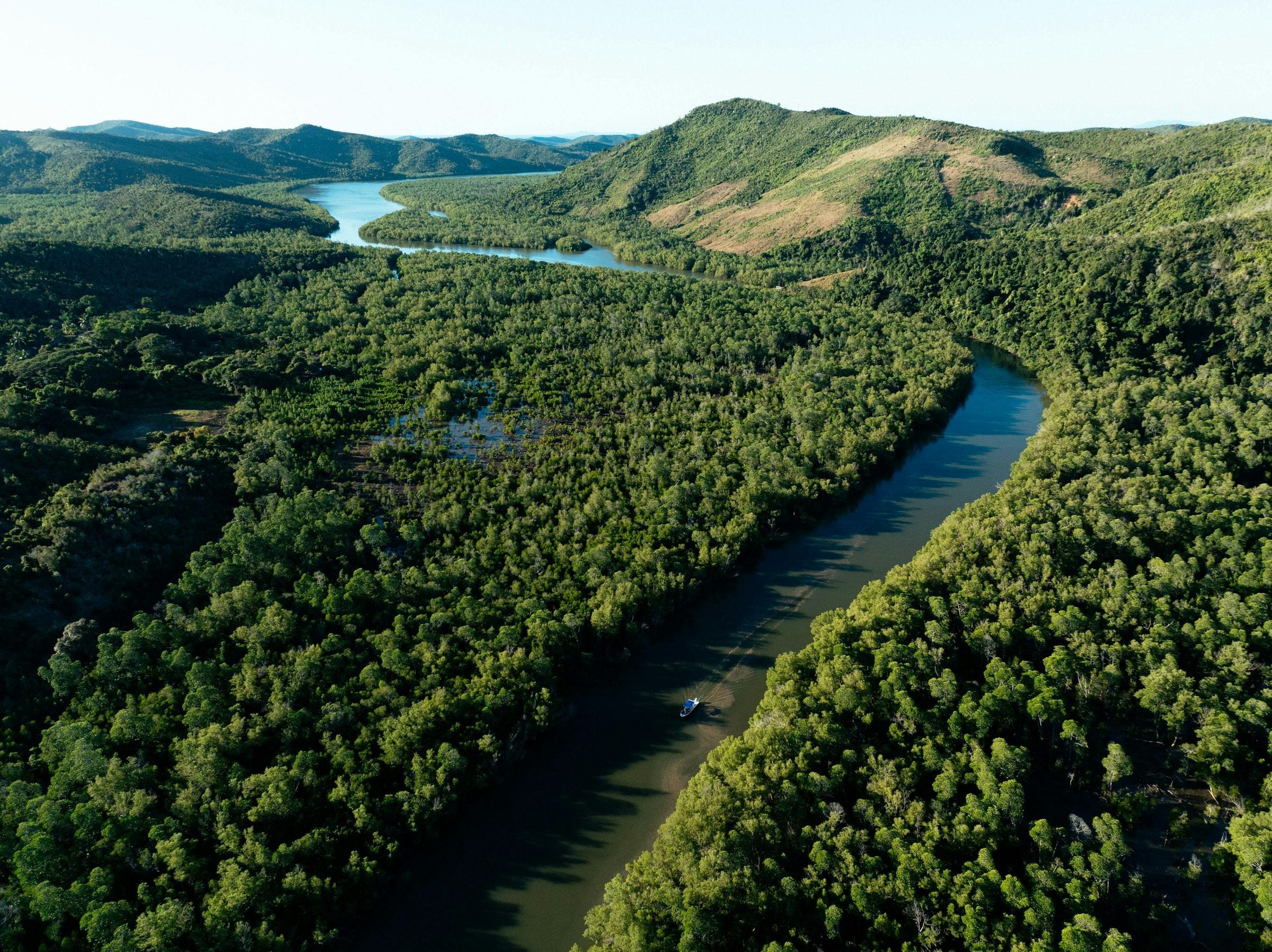 Aerial of a winding river threading through lush green forested hills, tiny boat below