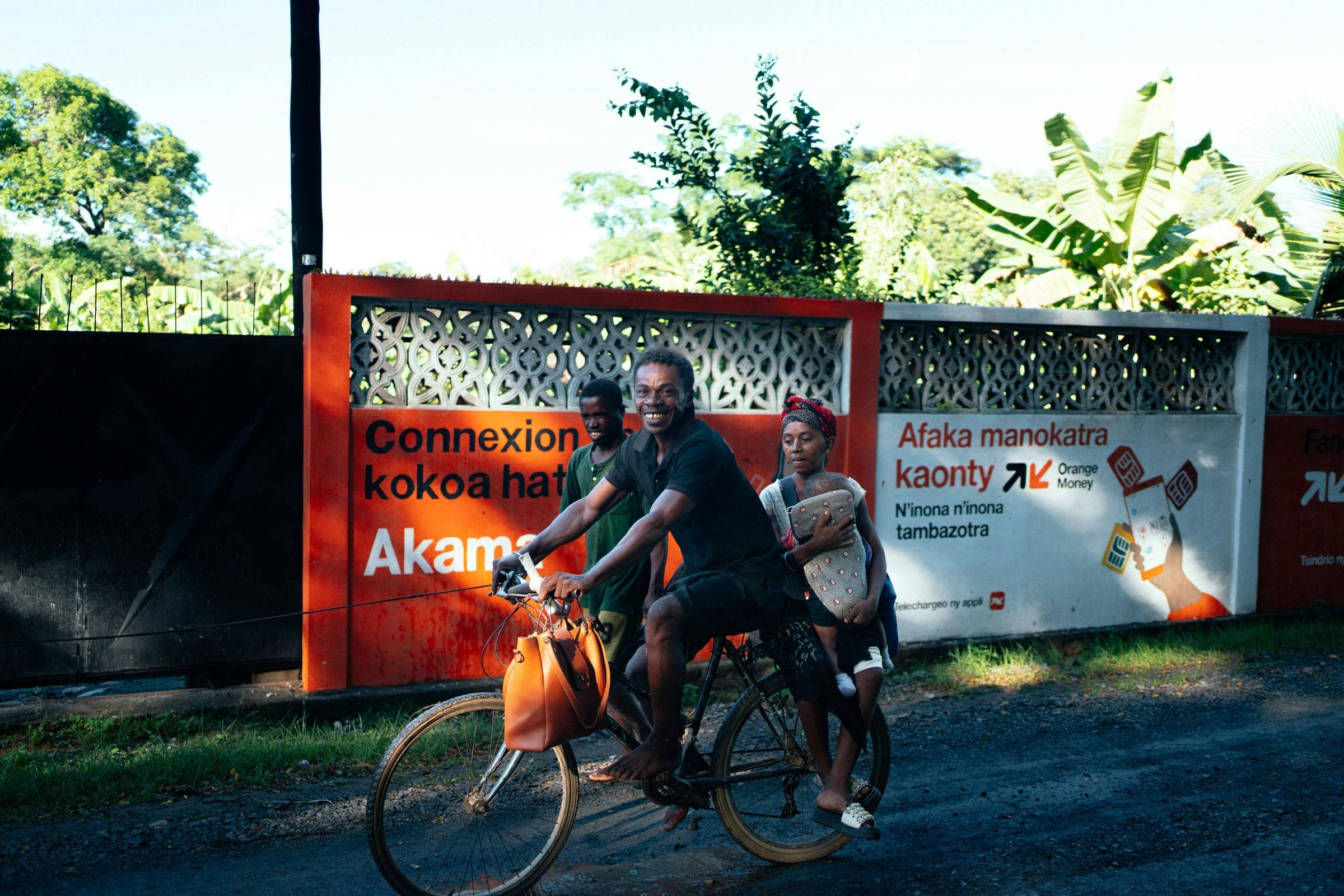 Smiling man cycling a dirt road with passengers past an Orange Money mural