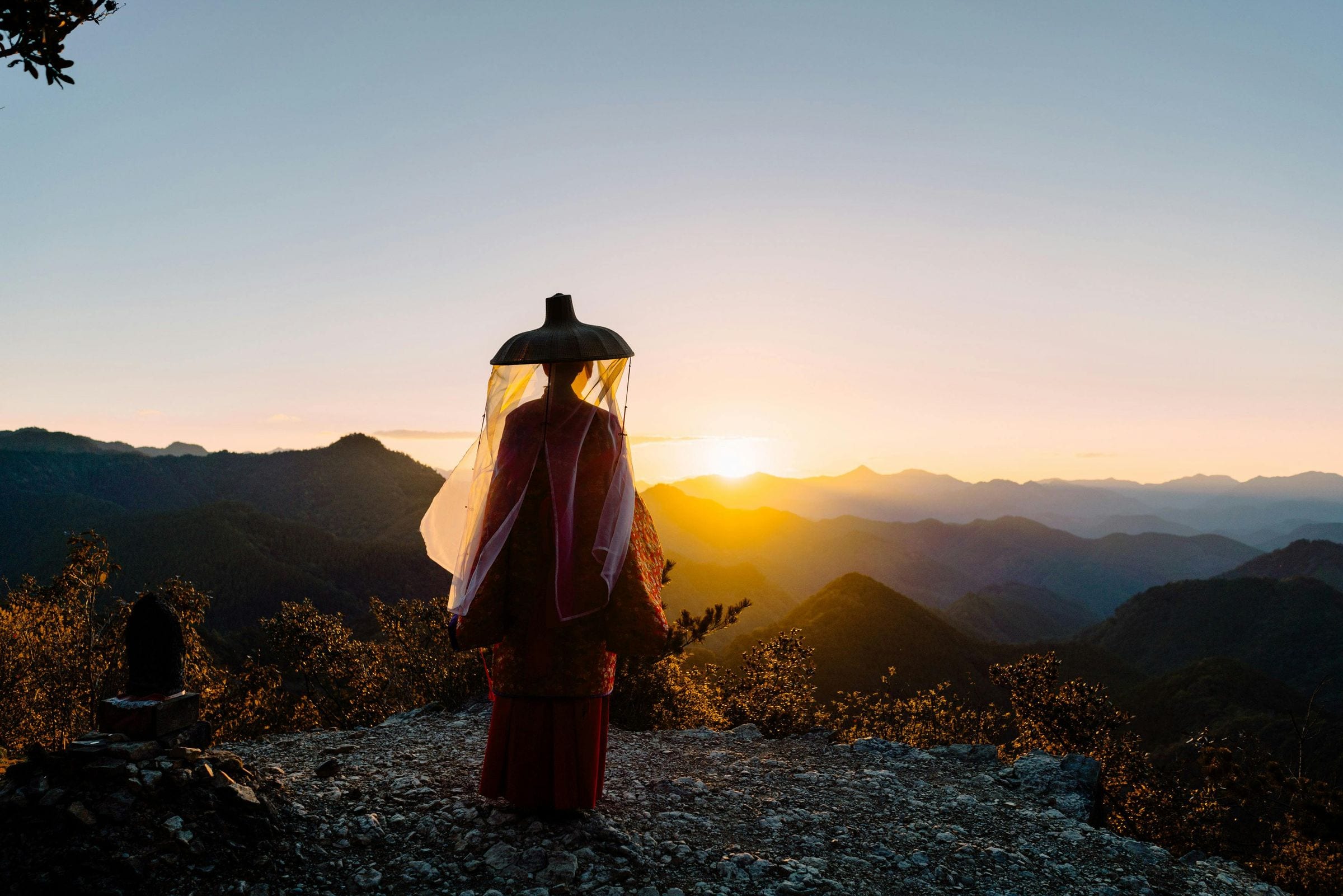 Figure in red robe and black lacquered hat facing sunrise over mountain ridges