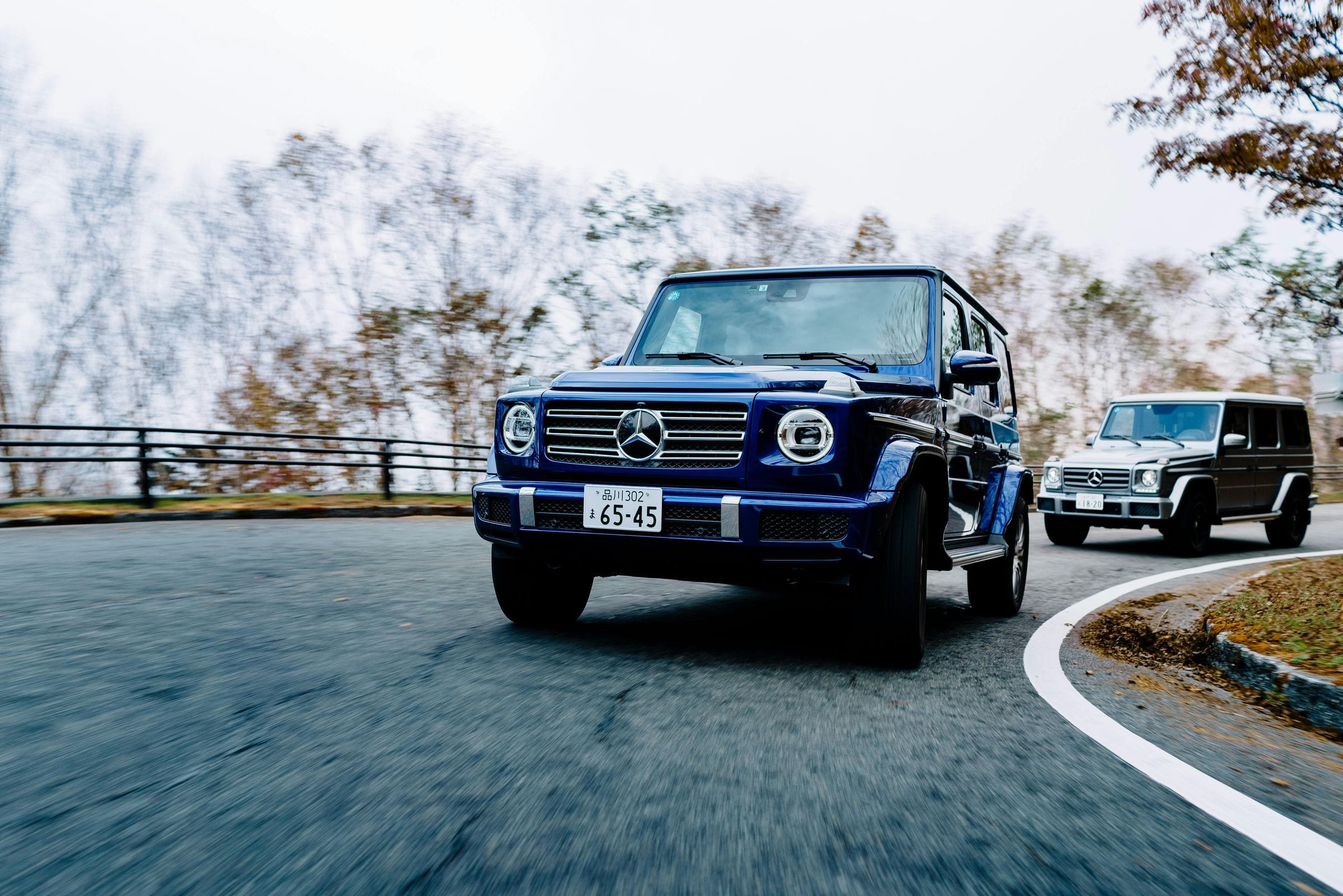 Blue Mercedes G-Class leading a black one around a wet mountain bend in autumn