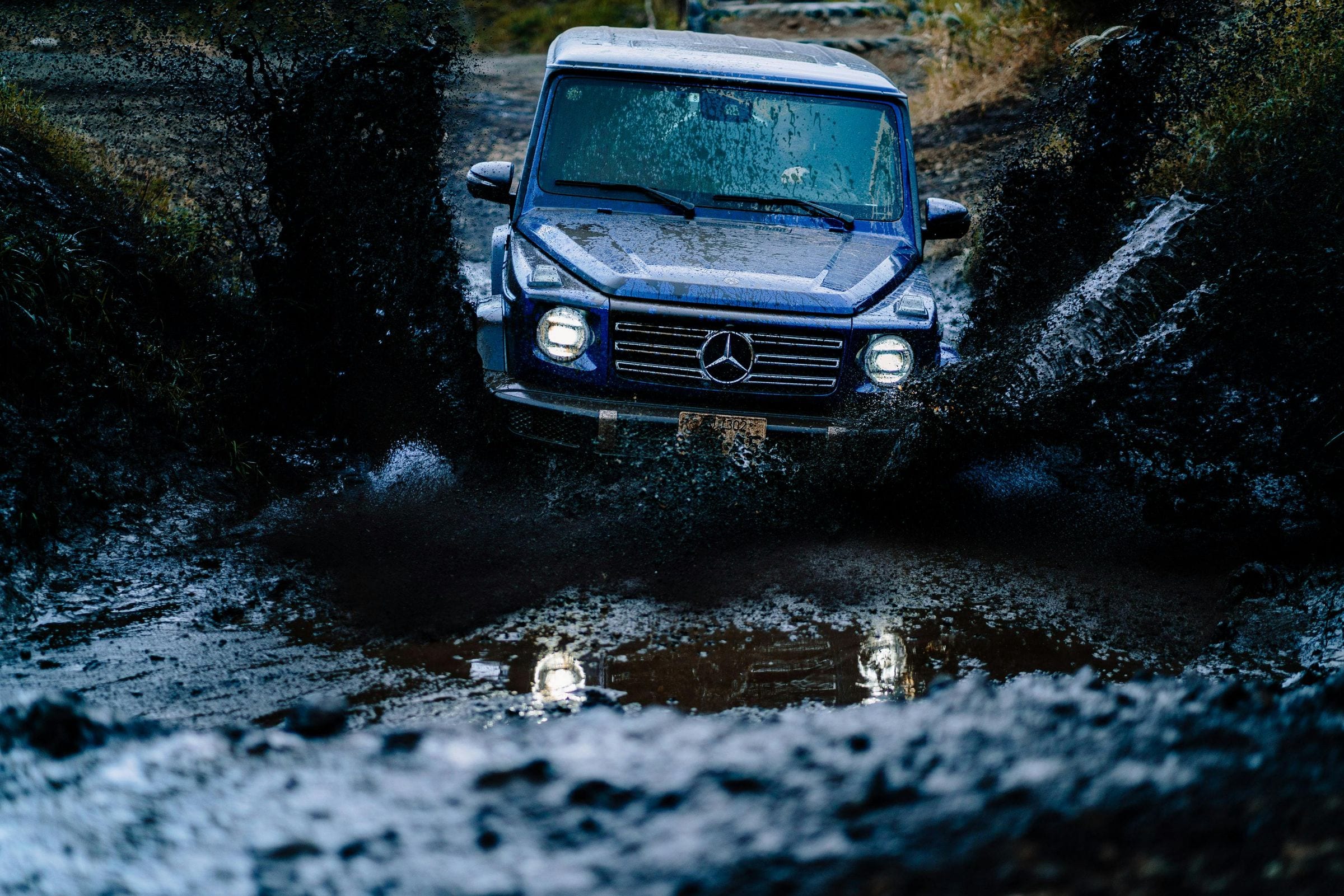 Blue Mercedes G-Class powering through a deep muddy off-road rut, headlights on