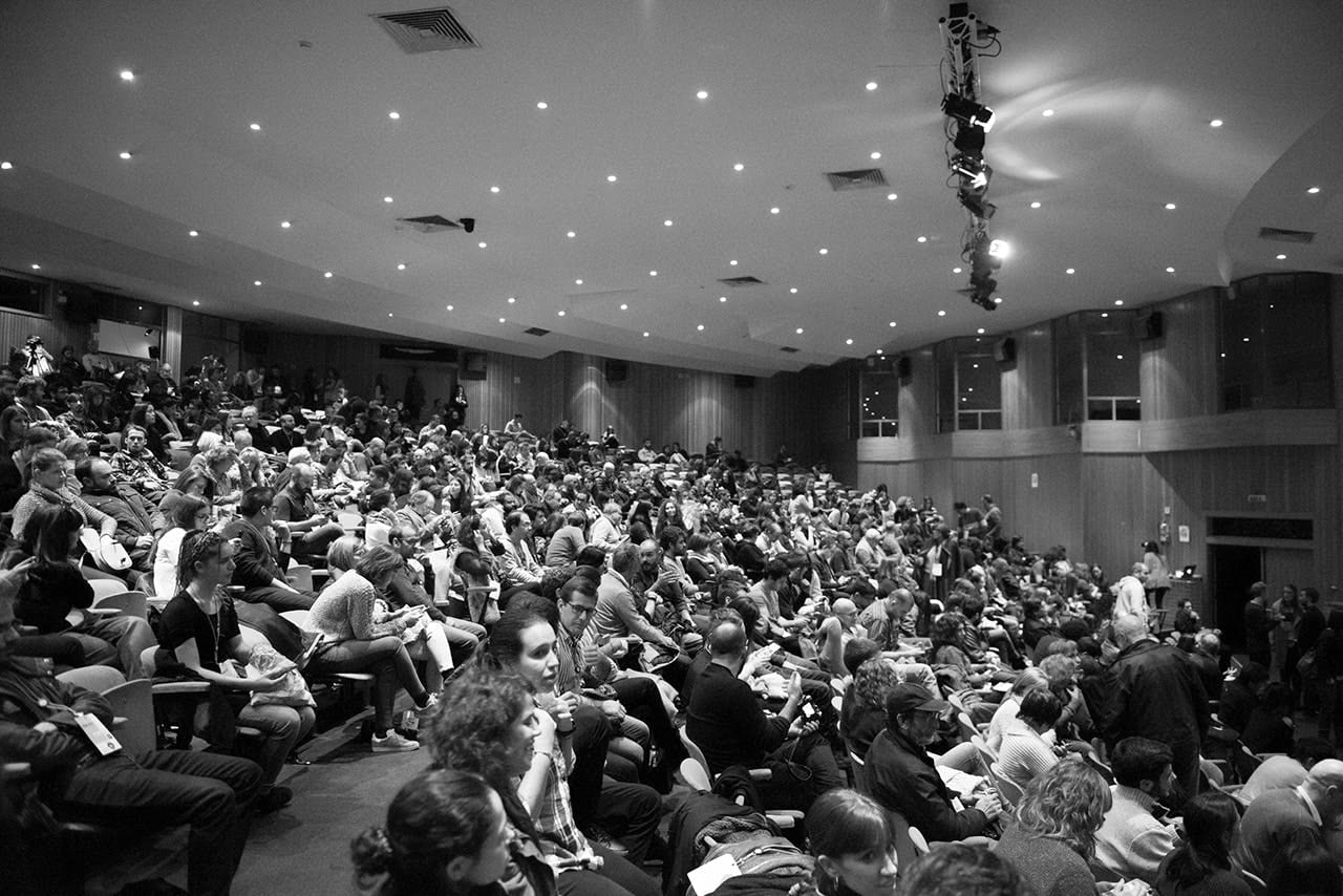 Packed auditorium of seated attendees at a talk, wide black-and-white view