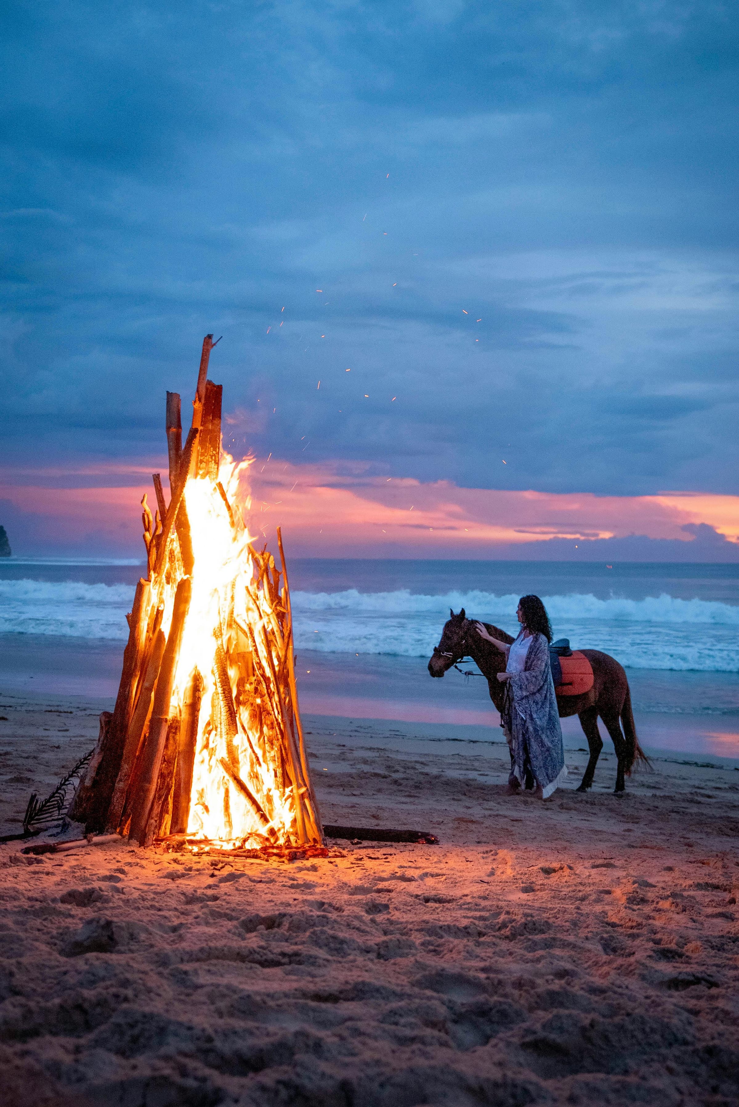 Woman with a saddled horse beside a towering driftwood bonfire on the beach at twilight