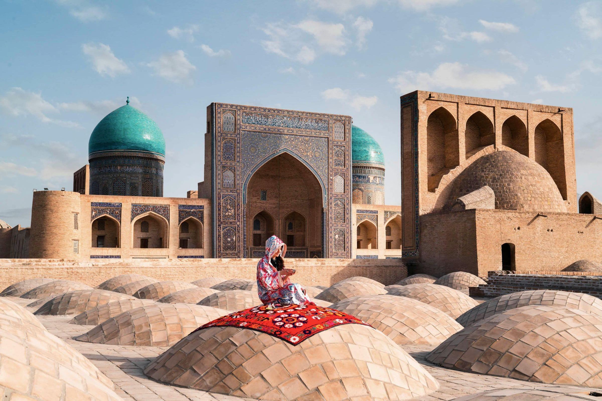 Woman in ikat robe on a red rug atop Bukhara's rooftop domes, tiled madrasa portal behind