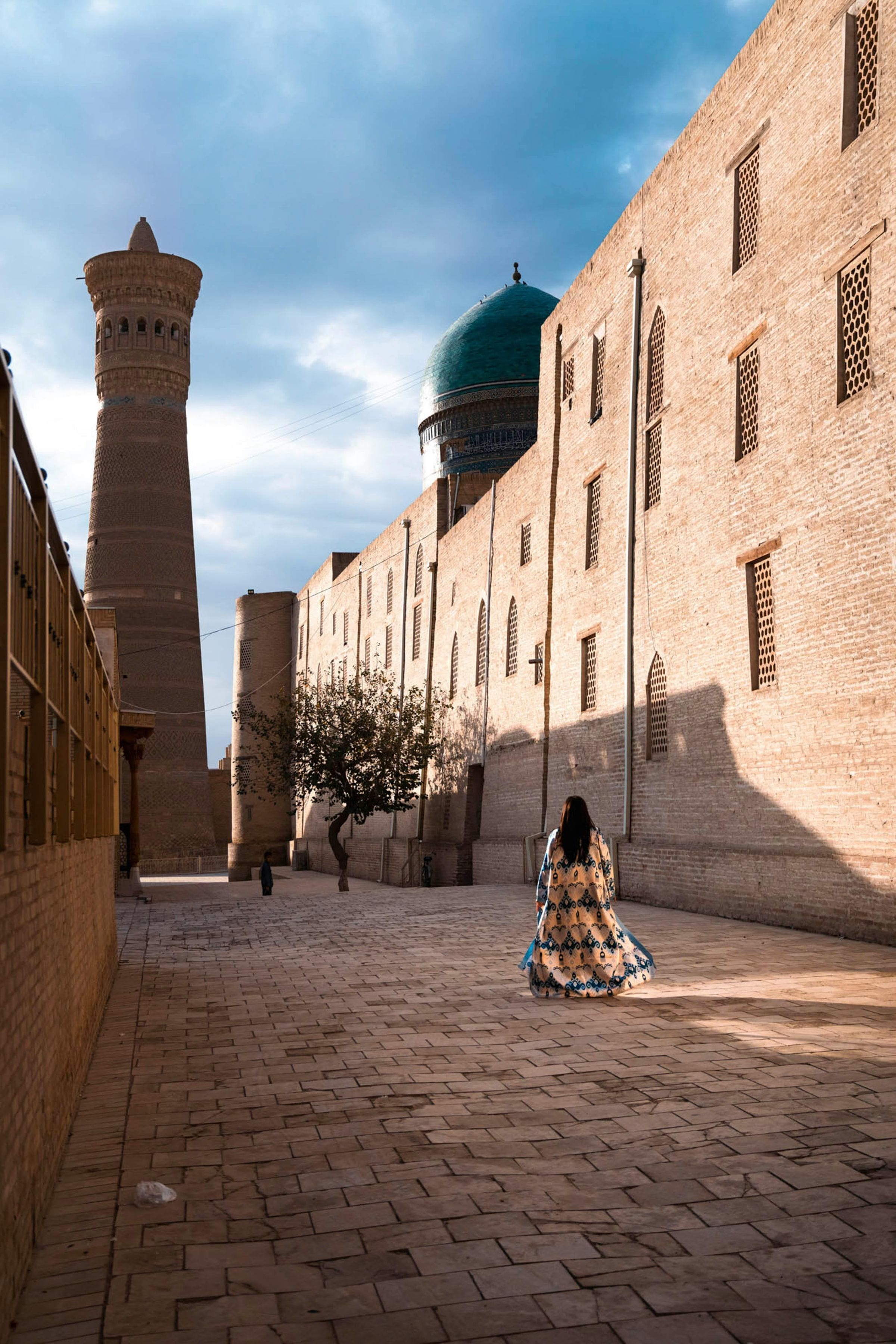 Woman in patterned robe walking an empty Bukhara lane toward the Kalyan minaret and turquoise dome