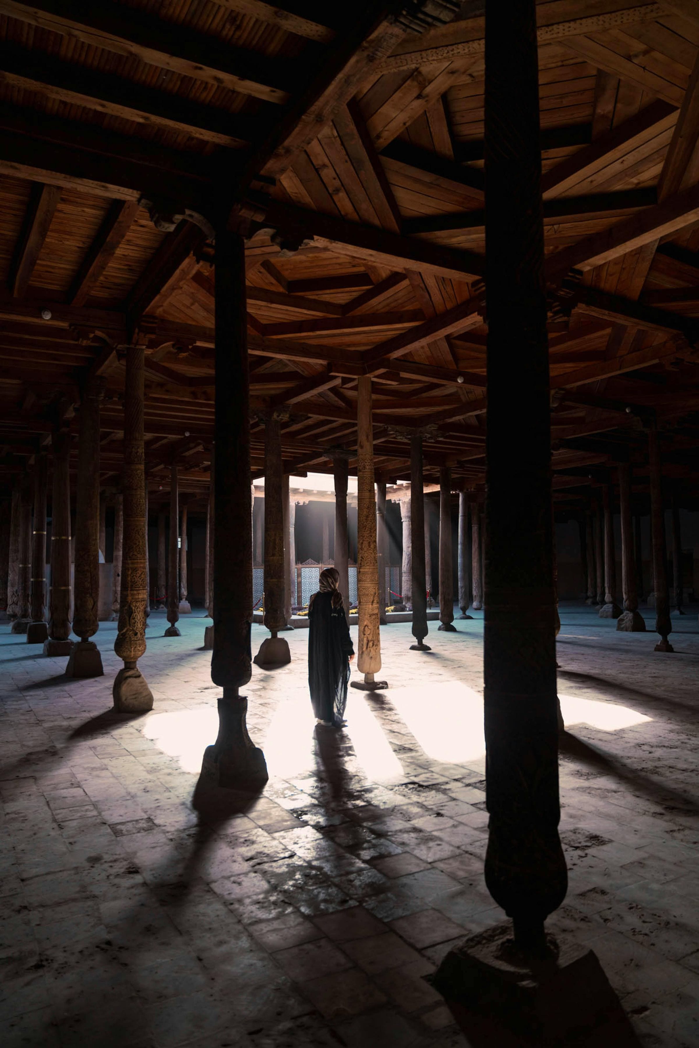 Lone figure among carved wooden columns of Khiva's Juma Mosque as light streams in