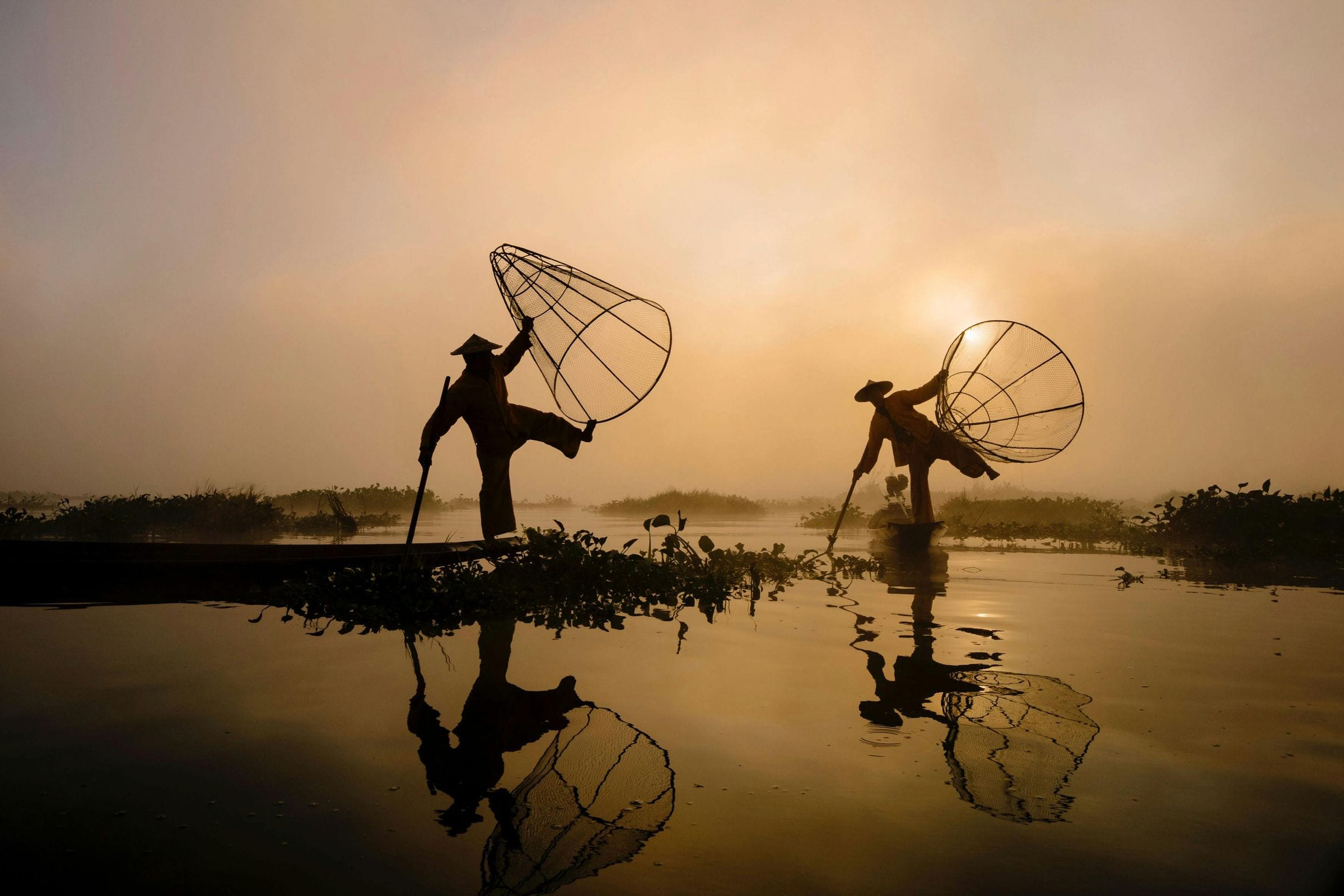 Two fishermen with conical nets silhouetted on a misty lake at golden dawn