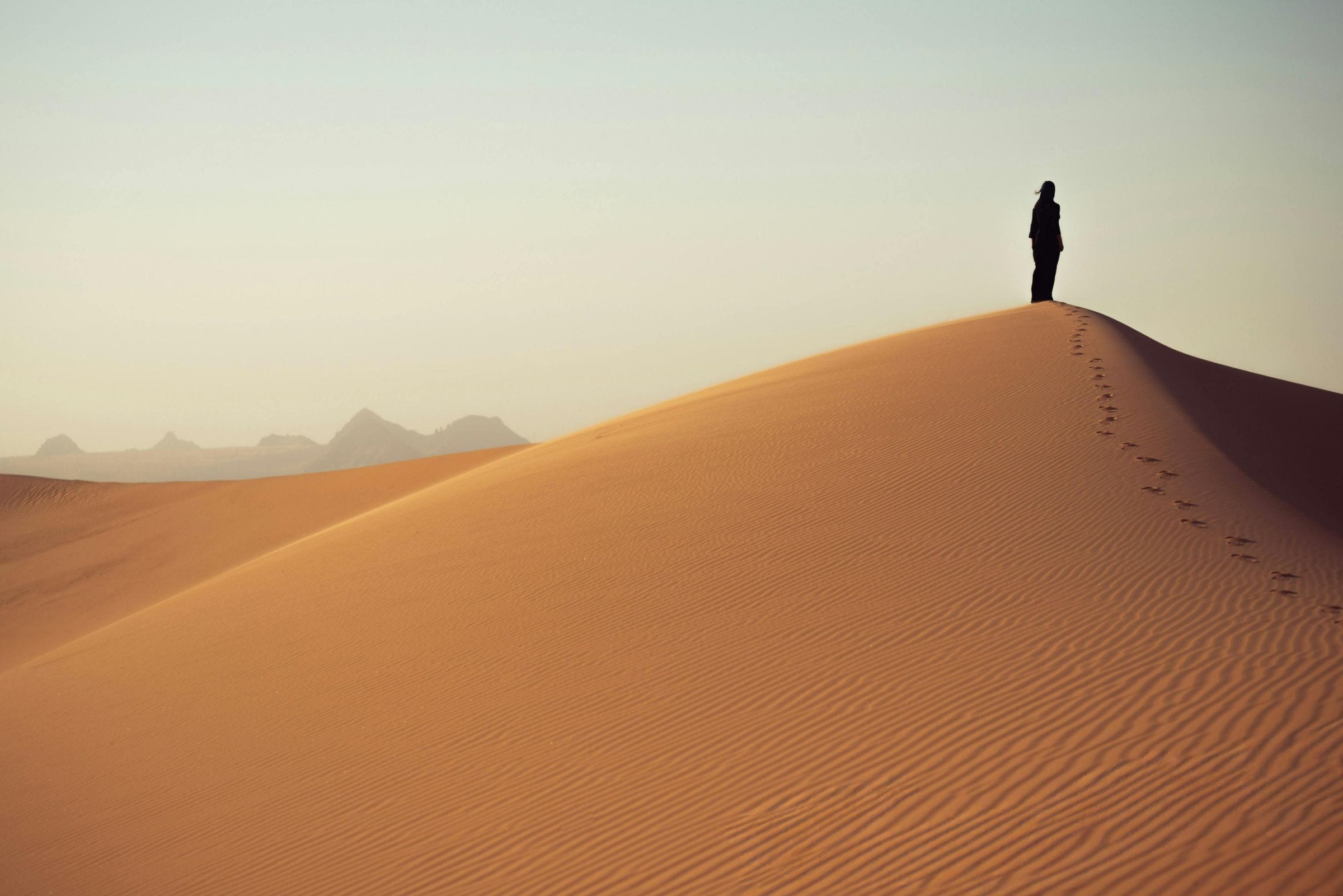 Lone figure on a golden sand dune ridge at dusk, footprints trailing behind