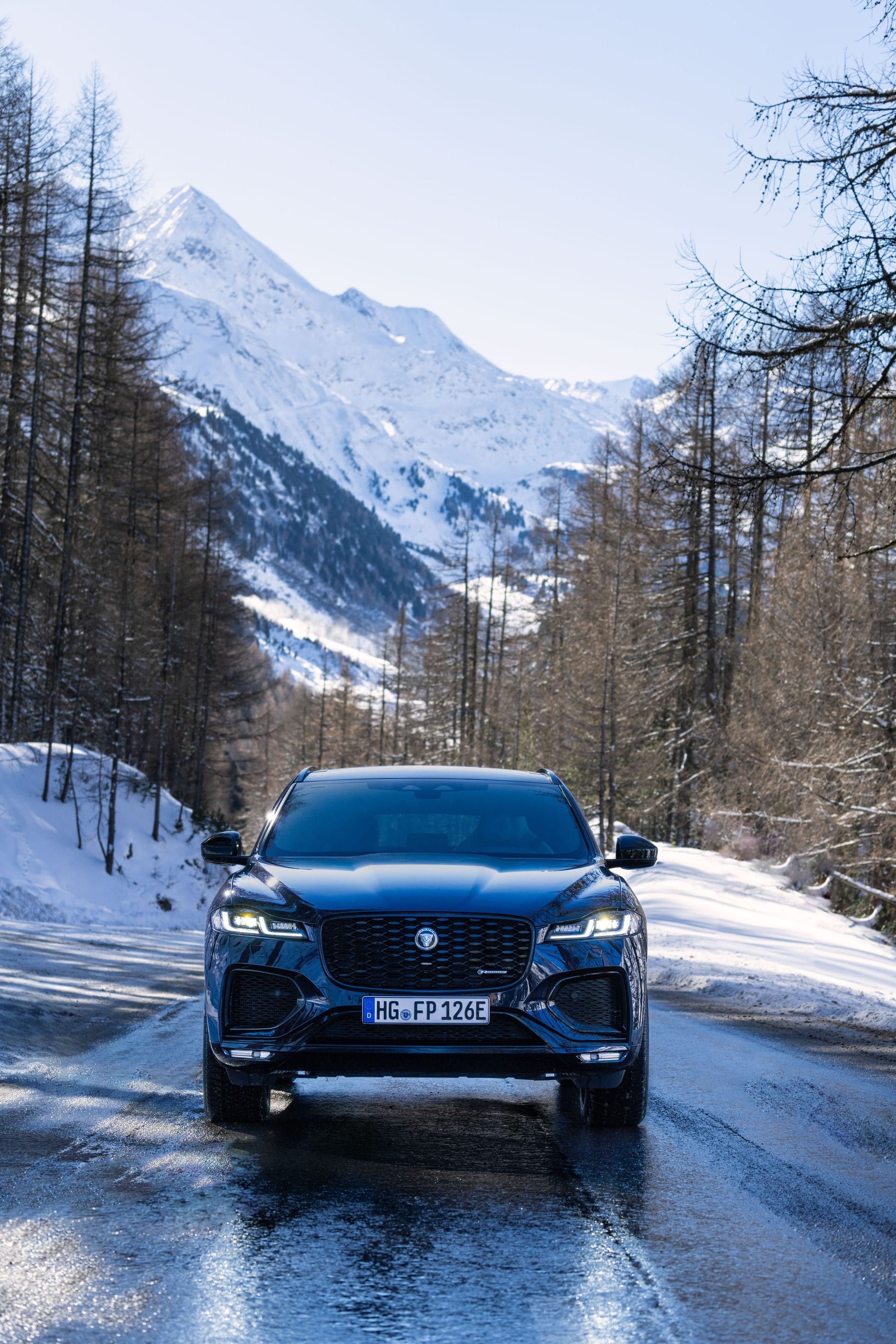 A navy Jaguar F-PACE on a wet mountain road framed by larches and a snowcapped peak in the Tirol