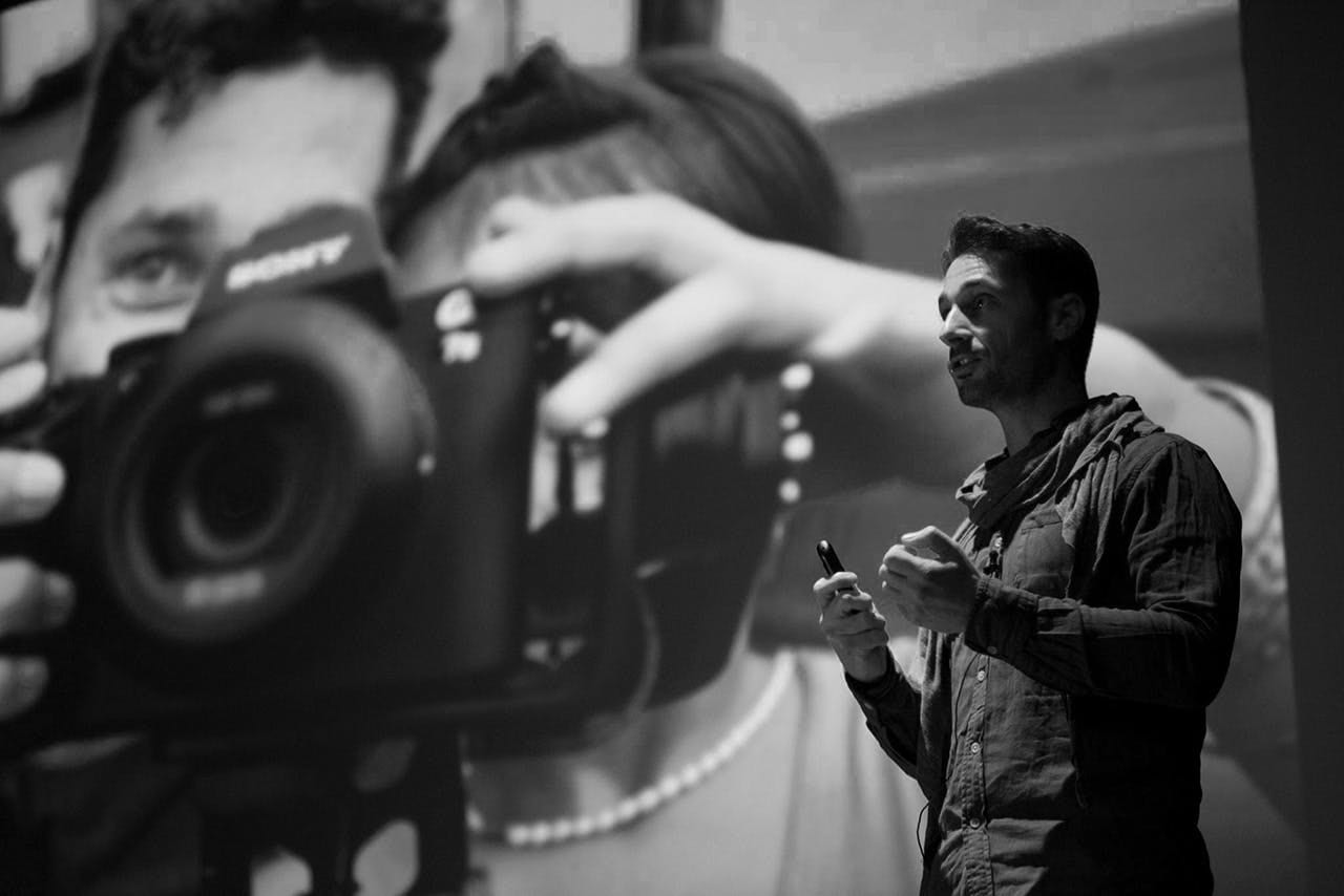 Speaker addressing a room beside a projected close-up of a Sony camera, black and white