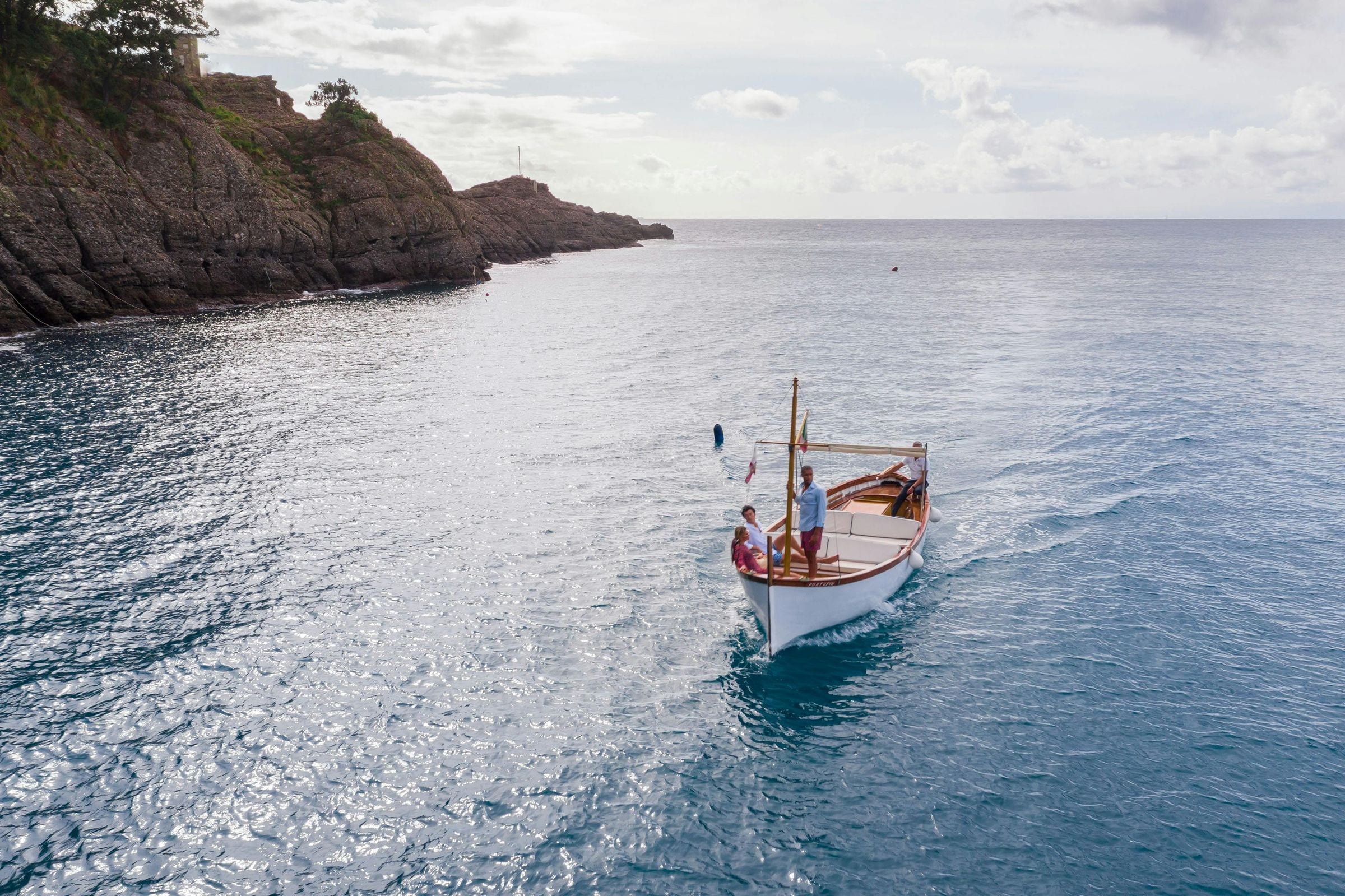 Classic wooden gozzo boat cruising past the Portofino cliffs under a cloudy sky