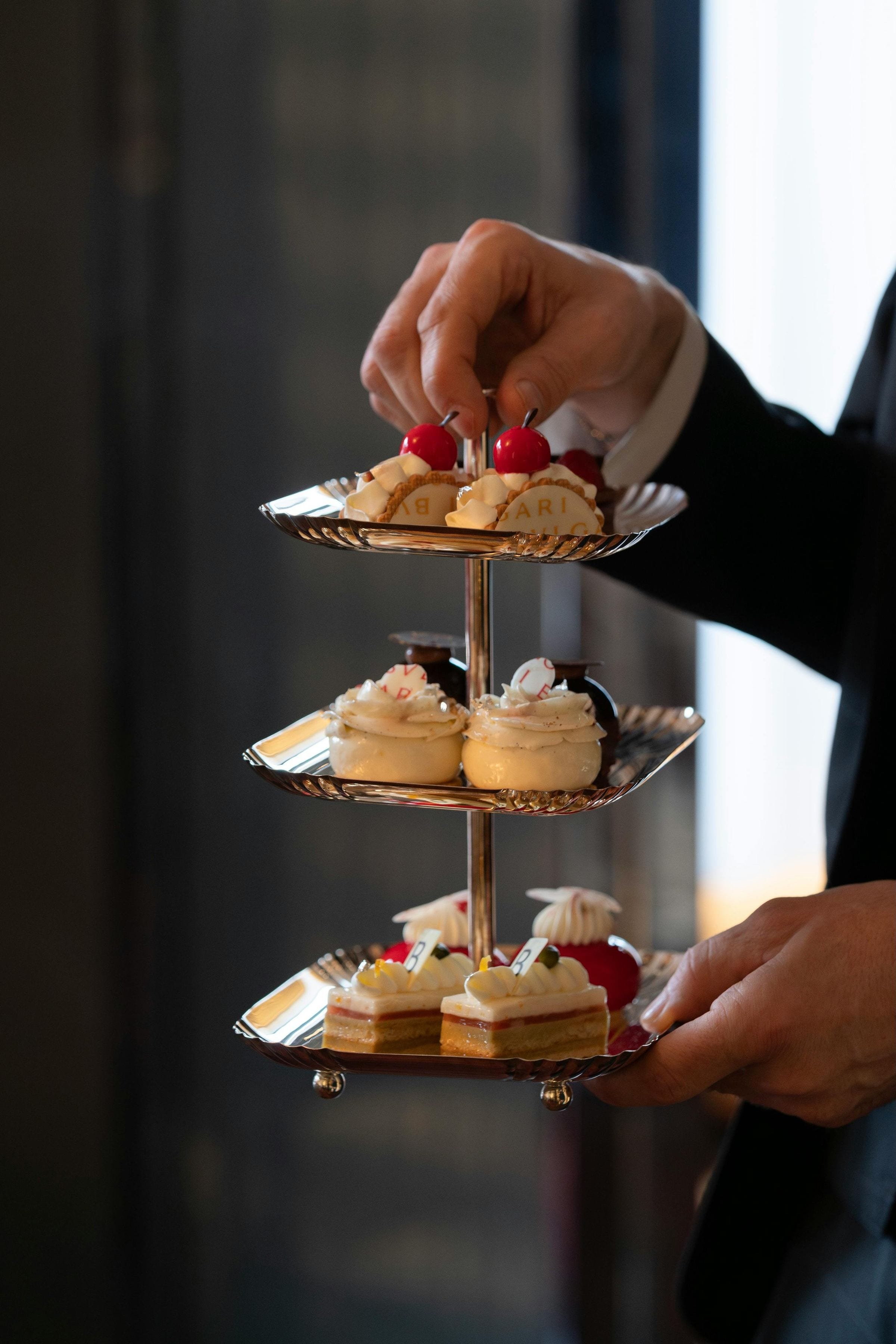 Server presenting a three-tier silver stand of Bvlgari-branded afternoon-tea pastries