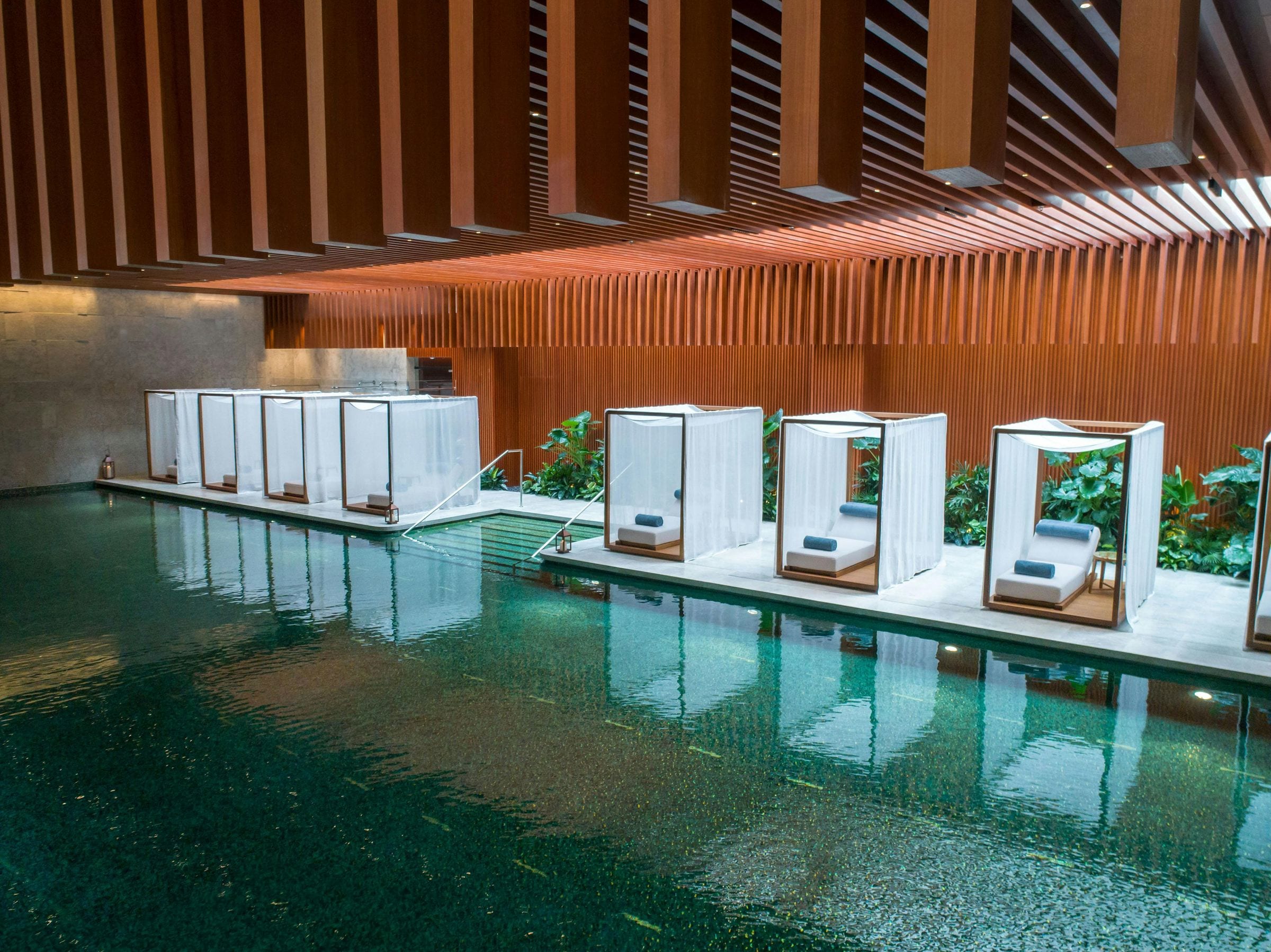 Indoor spa pool lined with white curtained daybed cabanas under a slatted timber ceiling