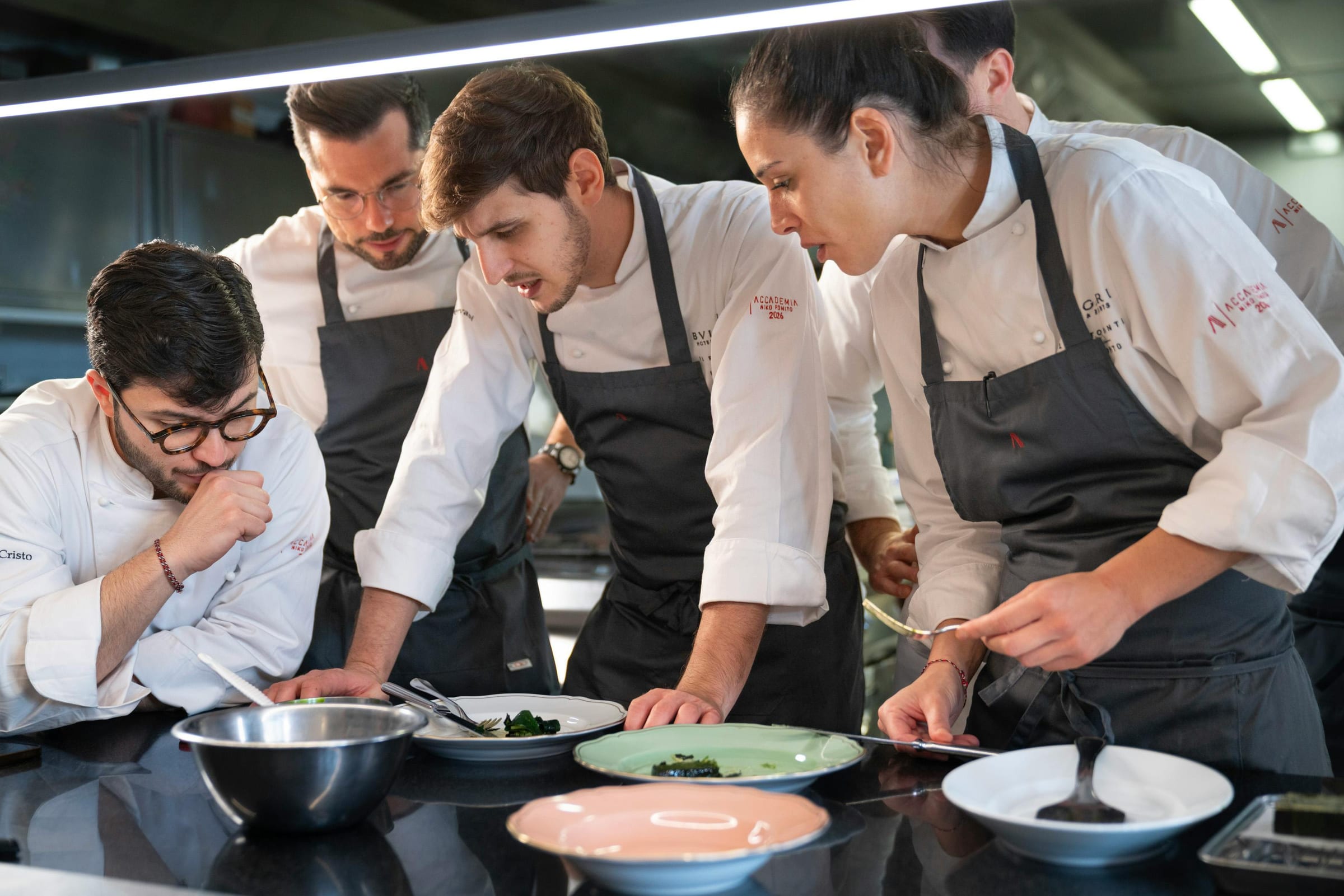 Five chefs in whites leaning over plated dishes at a stainless kitchen pass