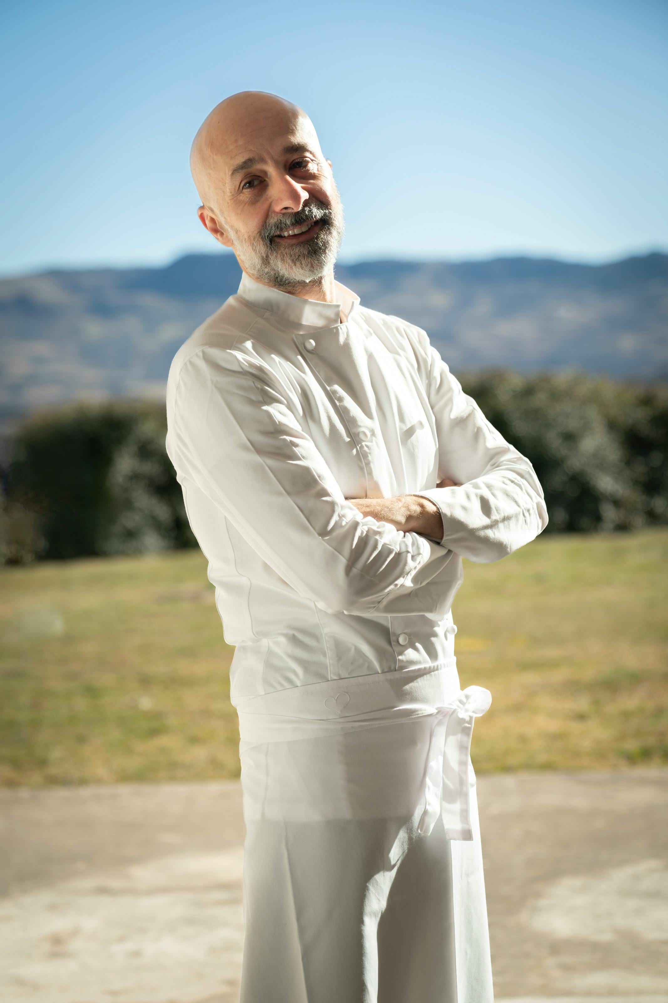 Smiling bearded chef in whites, arms crossed, against a sunny mountain backdrop