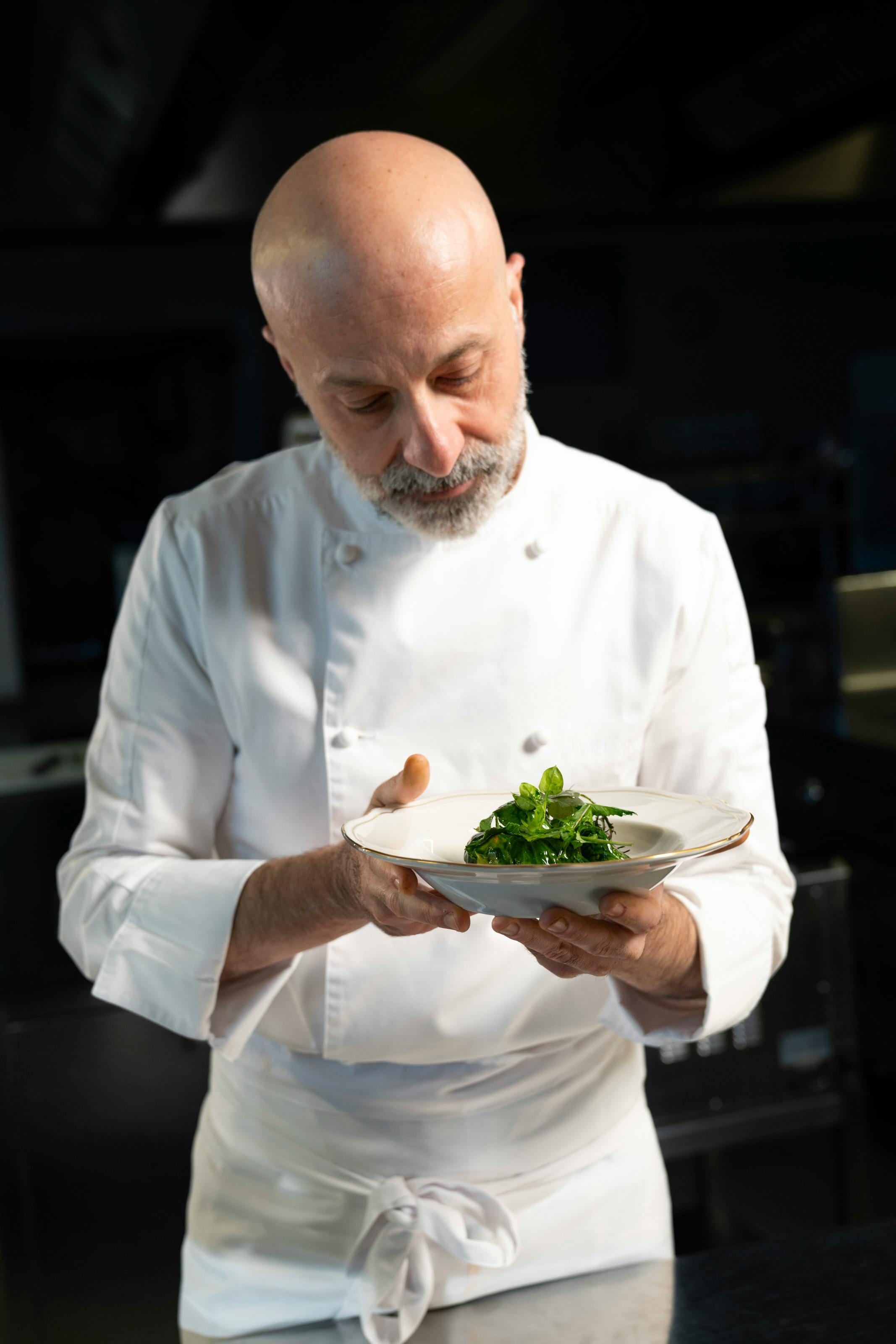 Bearded chef in whites studying a plated herb dish in a dark kitchen