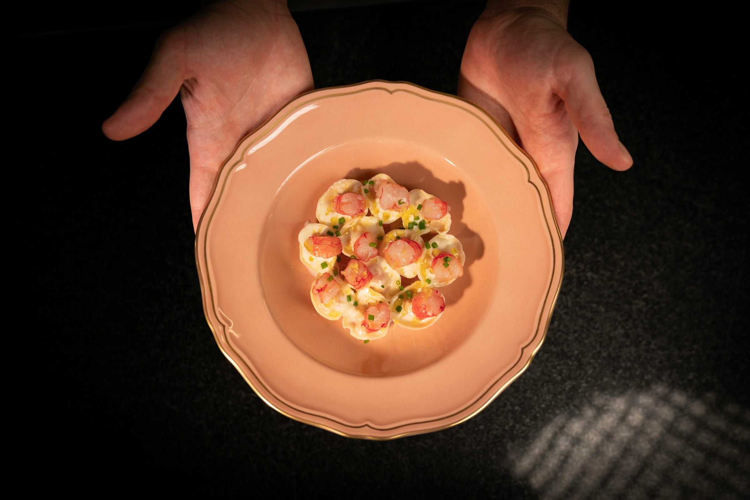 Hands presenting a blush plate of prawn tortelli with chives, seen from above