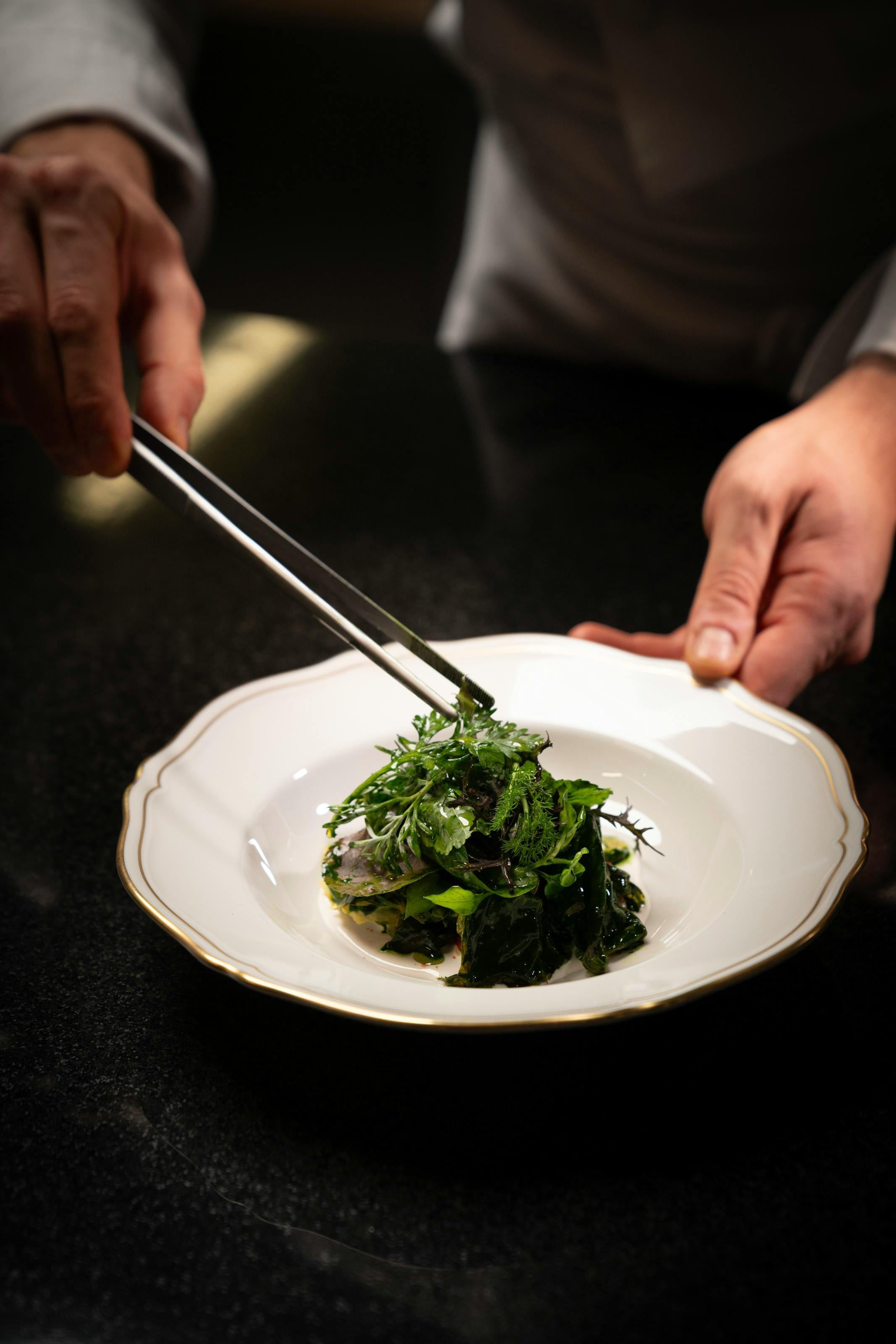 Chef lifting a fresh herb garnish onto a gold-rimmed plate with tongs
