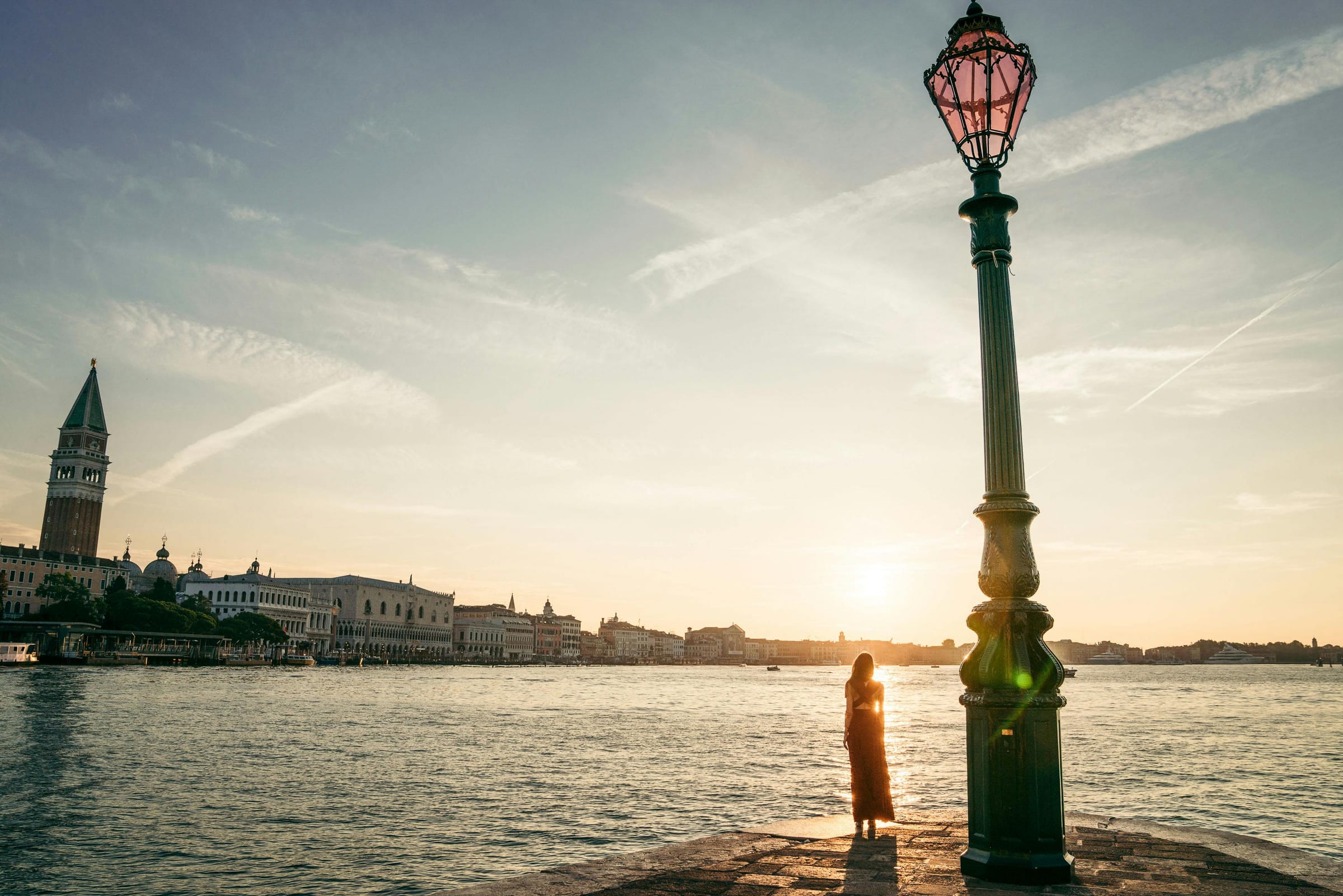 Woman silhouetted by a Venetian lamp post at sunrise, San Marco campanile across the water