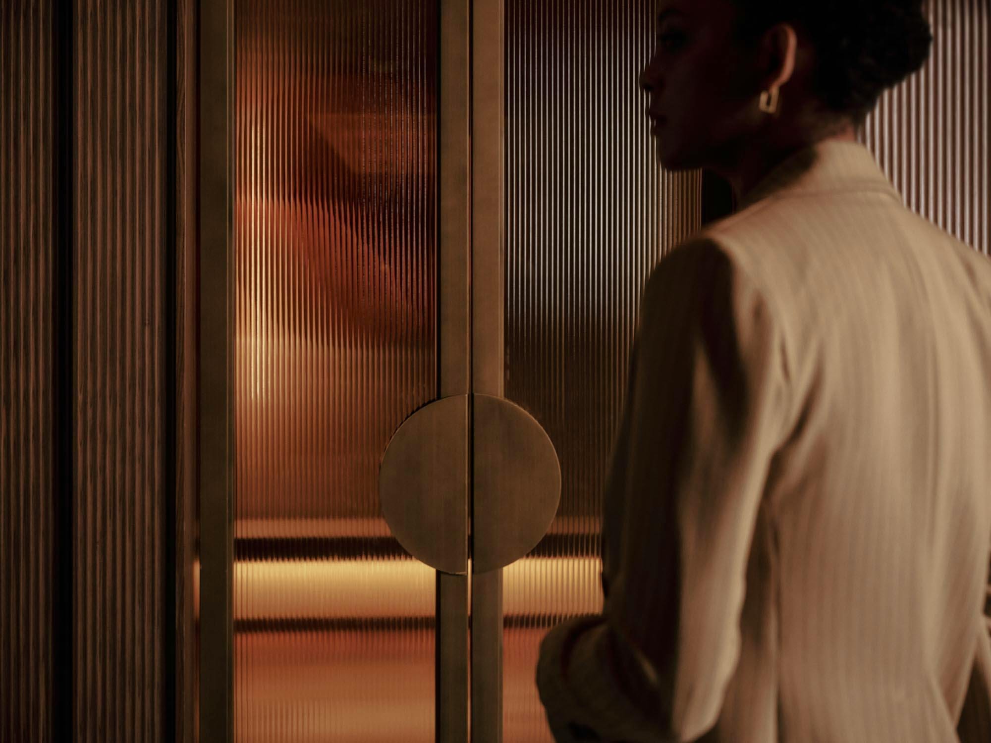 Woman in cream silhouetted beside a fluted-glass and brass door in warm interior light