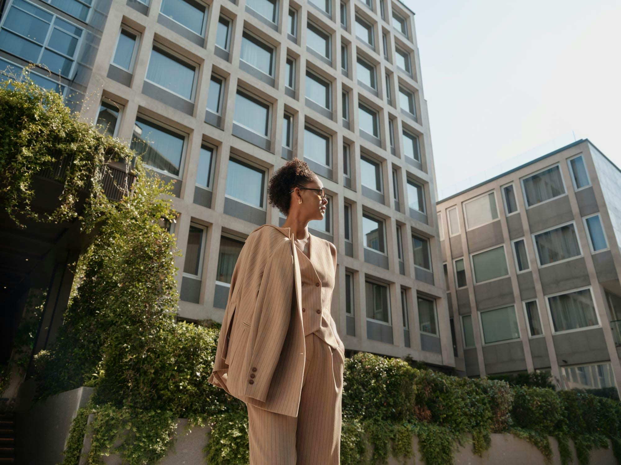 Woman in a beige pinstripe suit before Casa Brera's gridded façade and cascading greenery