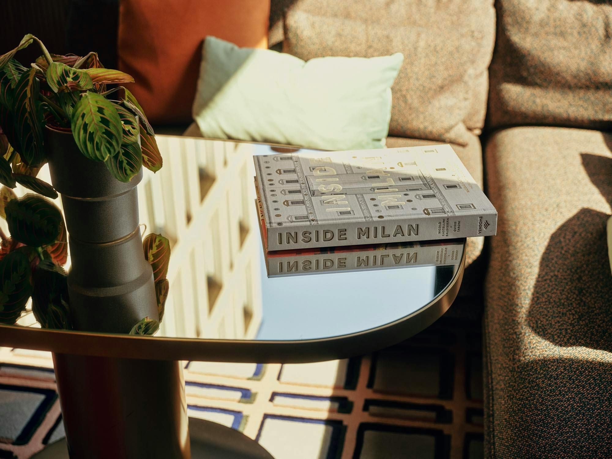Inside Milan coffee-table books stacked on a mirrored side table beside a leafy plant