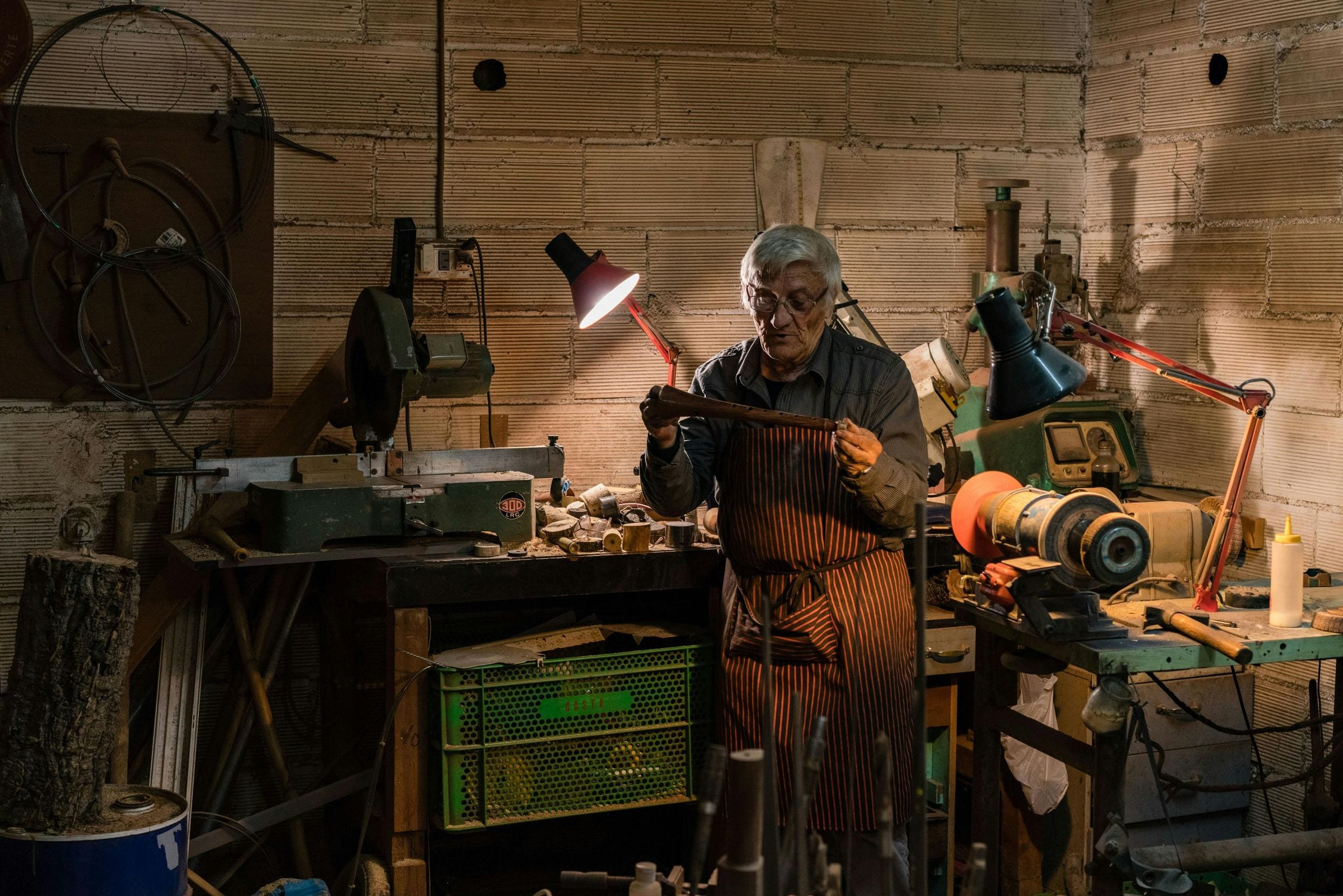 Elderly craftsman works at a lathe under a red lamp in a cluttered workshop