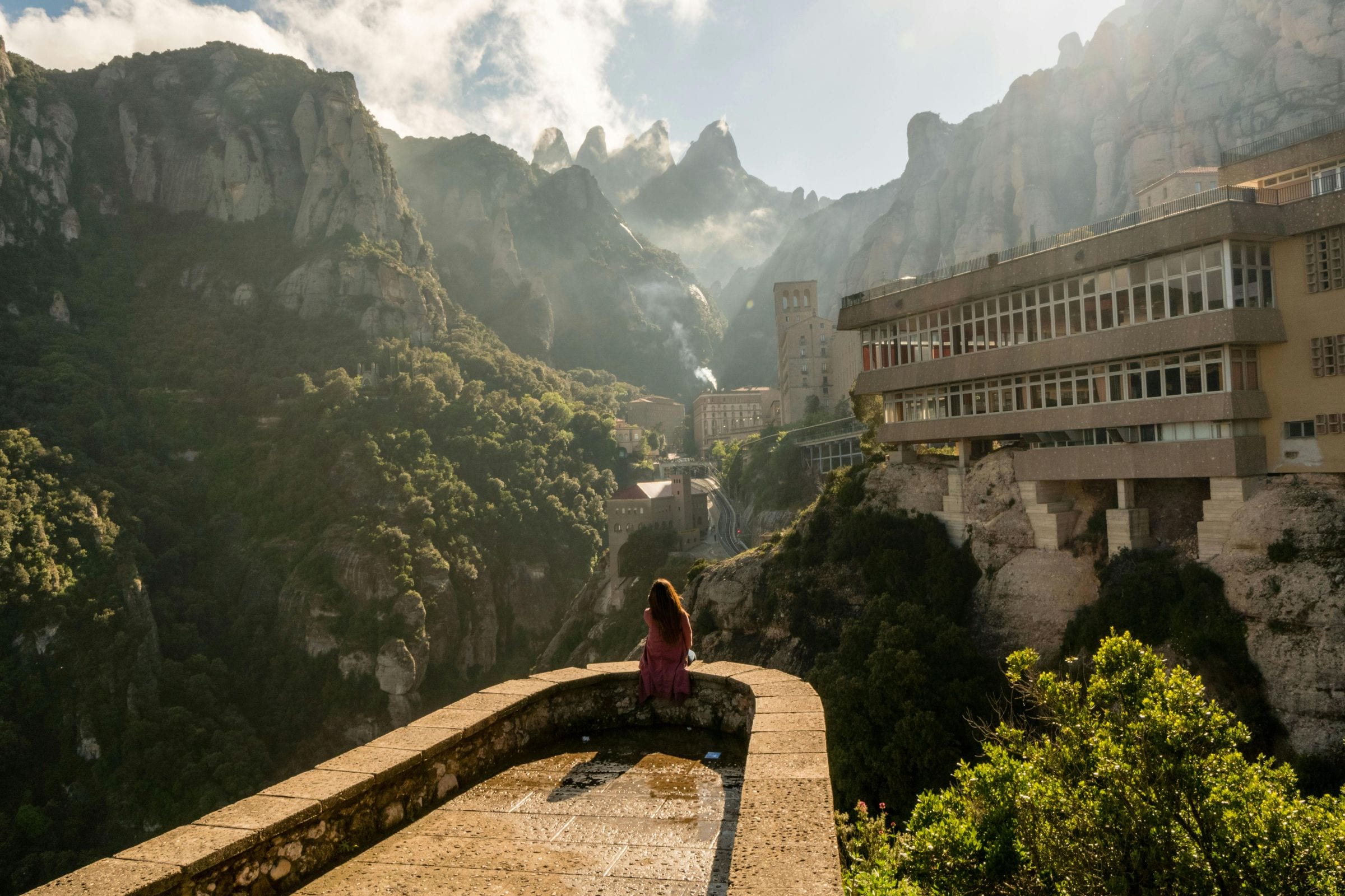 Woman in pink sits on a stone viewpoint facing Montserrat's jagged peaks and monastery