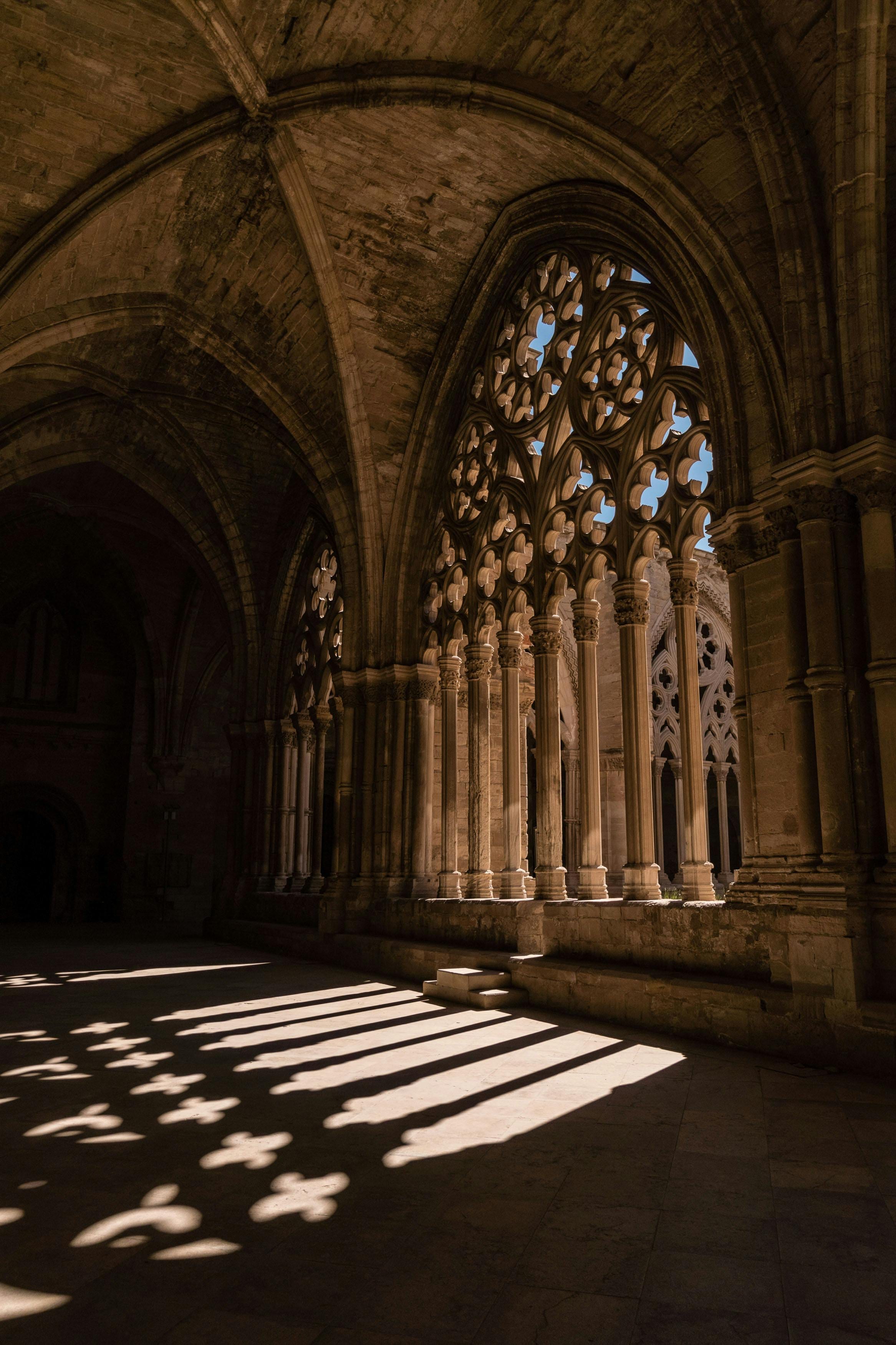 Sunlight through Gothic cloister tracery casting lace-like shadows on the stone floor