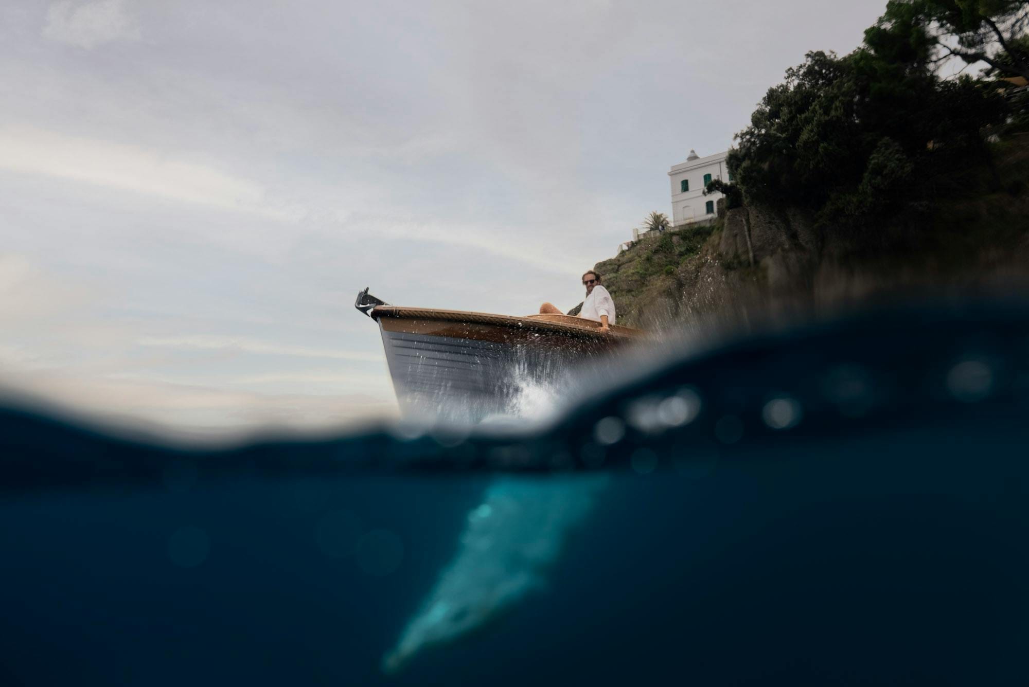 Half-underwater view of a wooden boat's bow, man aboard beside a cliffside villa