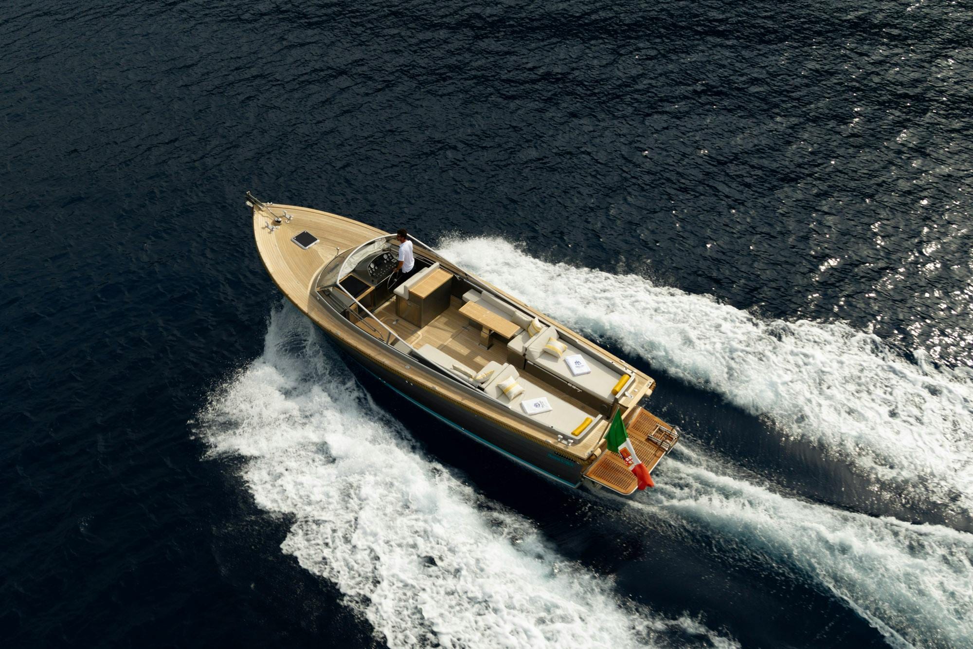 Wooden Mussini boat speeding across deep-blue water leaving a white wake, aerial view
