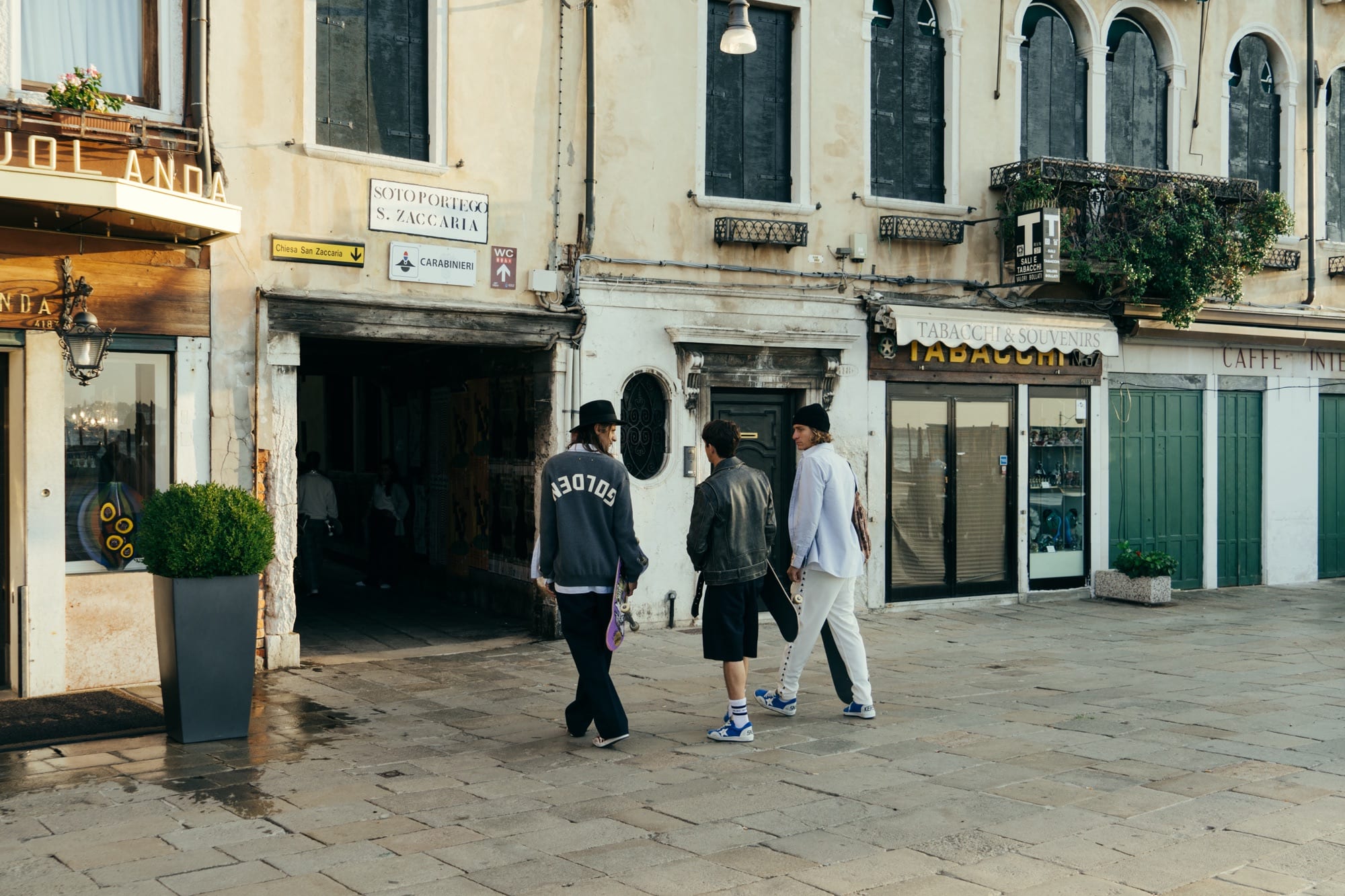 Three skaters carrying their boards through a Venetian campo at golden hour, wearing Golden Goose