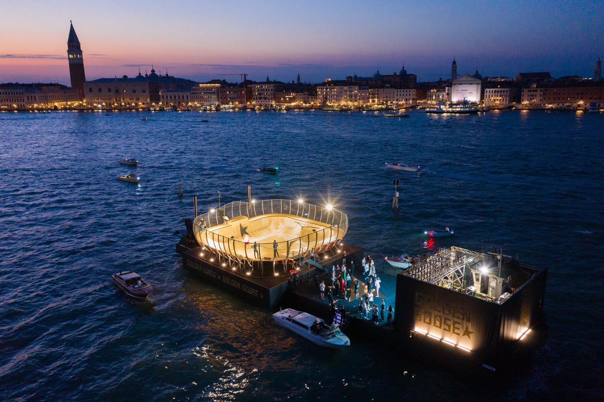 Aerial view at dusk of the illuminated Golden Goose skate bowl floating on the Venetian Lagoon, with the Venice skyline behind