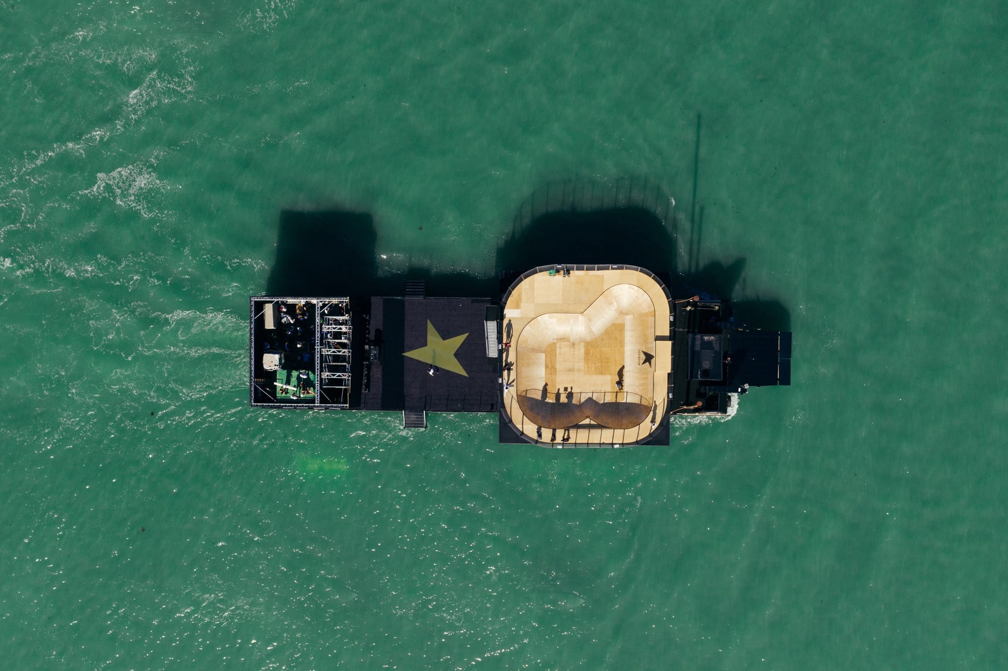 Top-down aerial of the black barge carrying the wooden skate bowl and a gold star on the green lagoon water