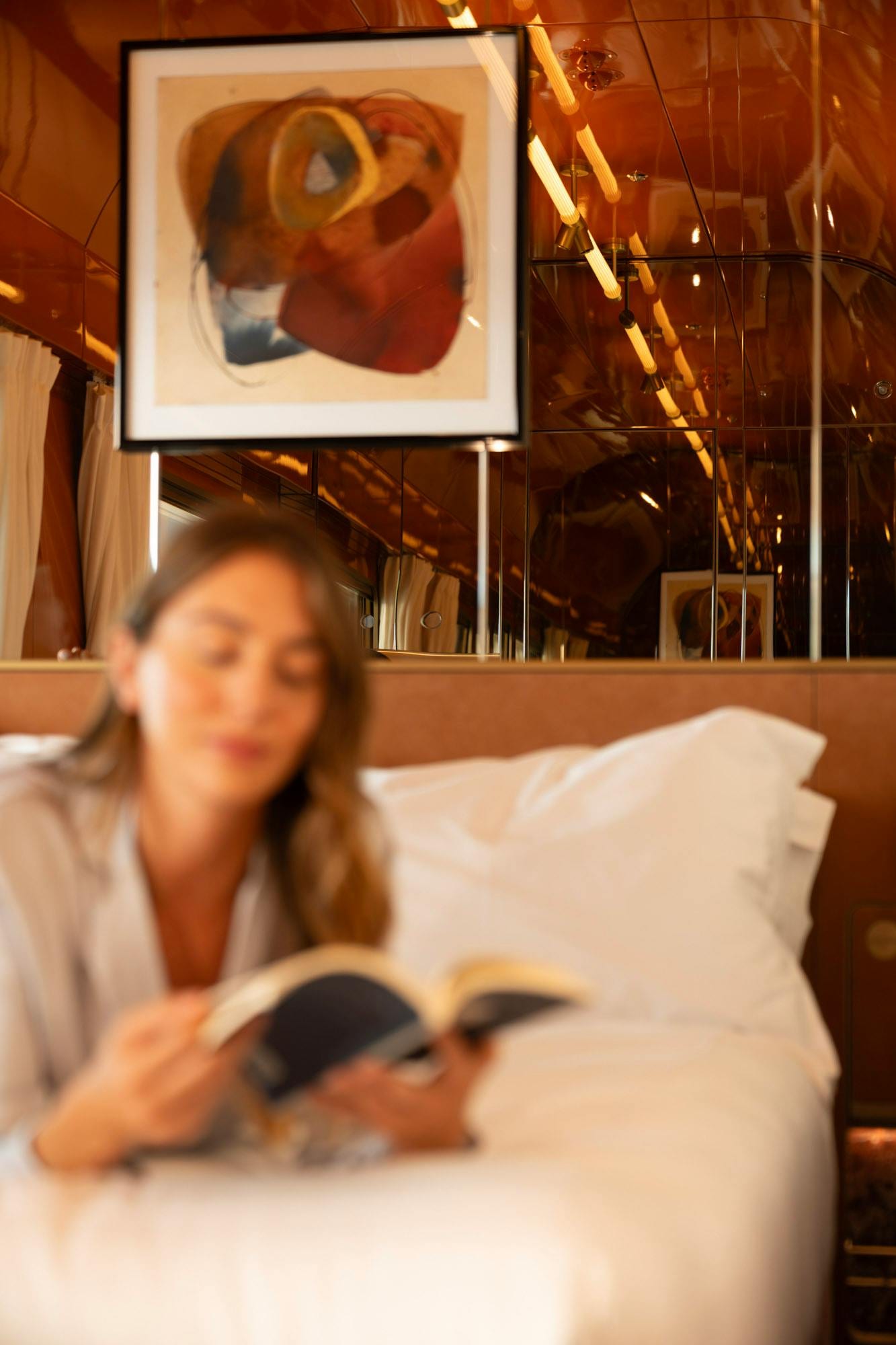 Woman reading in bed inside a warm mirrored train suite with framed abstract art