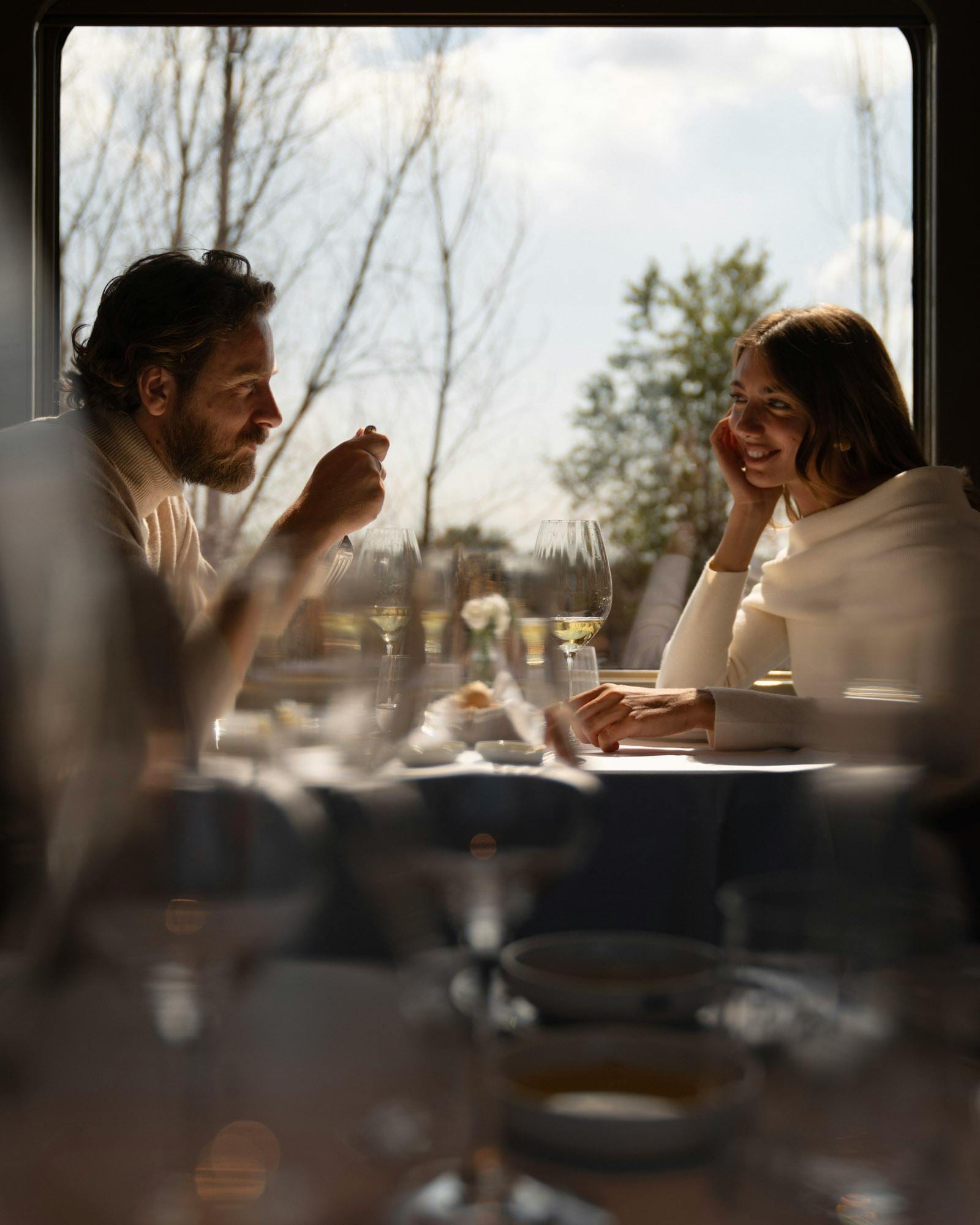 Couple dining at a train window table with white wine, backlit by passing countryside