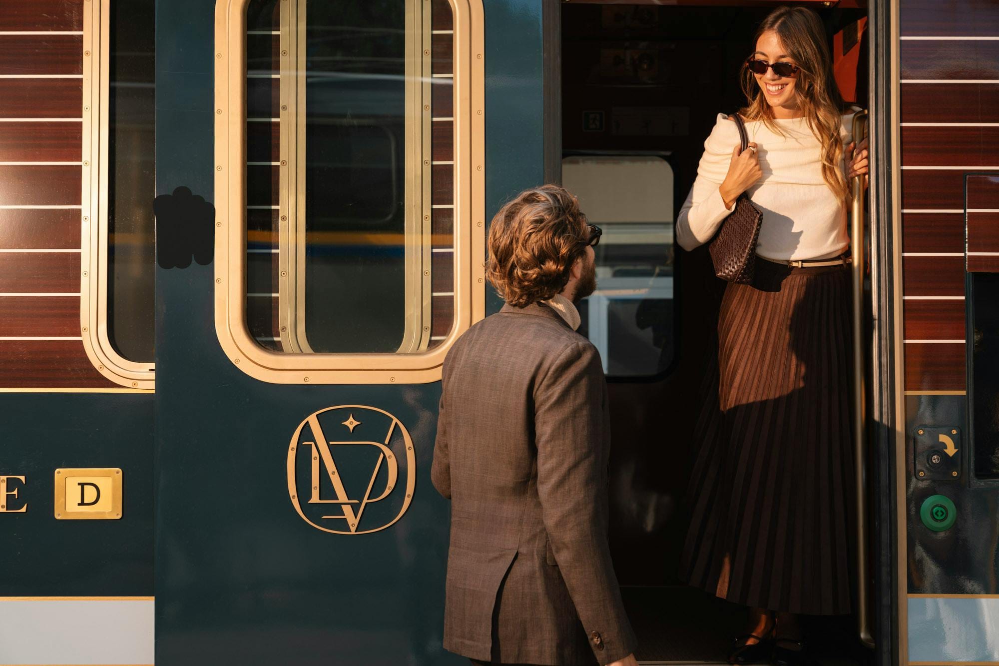 Man in a grey suit greets a smiling woman boarding the train at golden hour