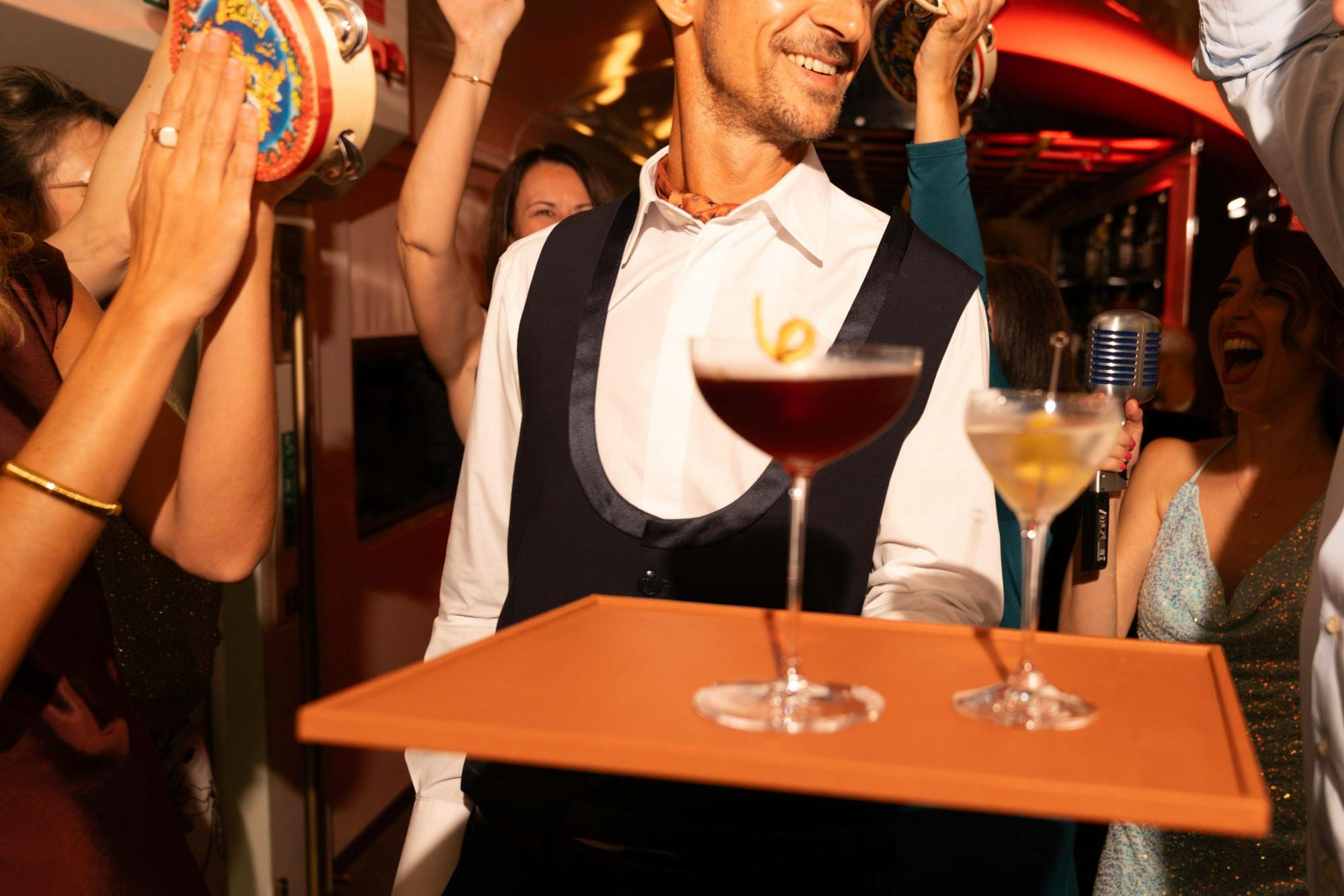Waiter carries a cocktail tray through a lively carriage as guests sing and play tambourines