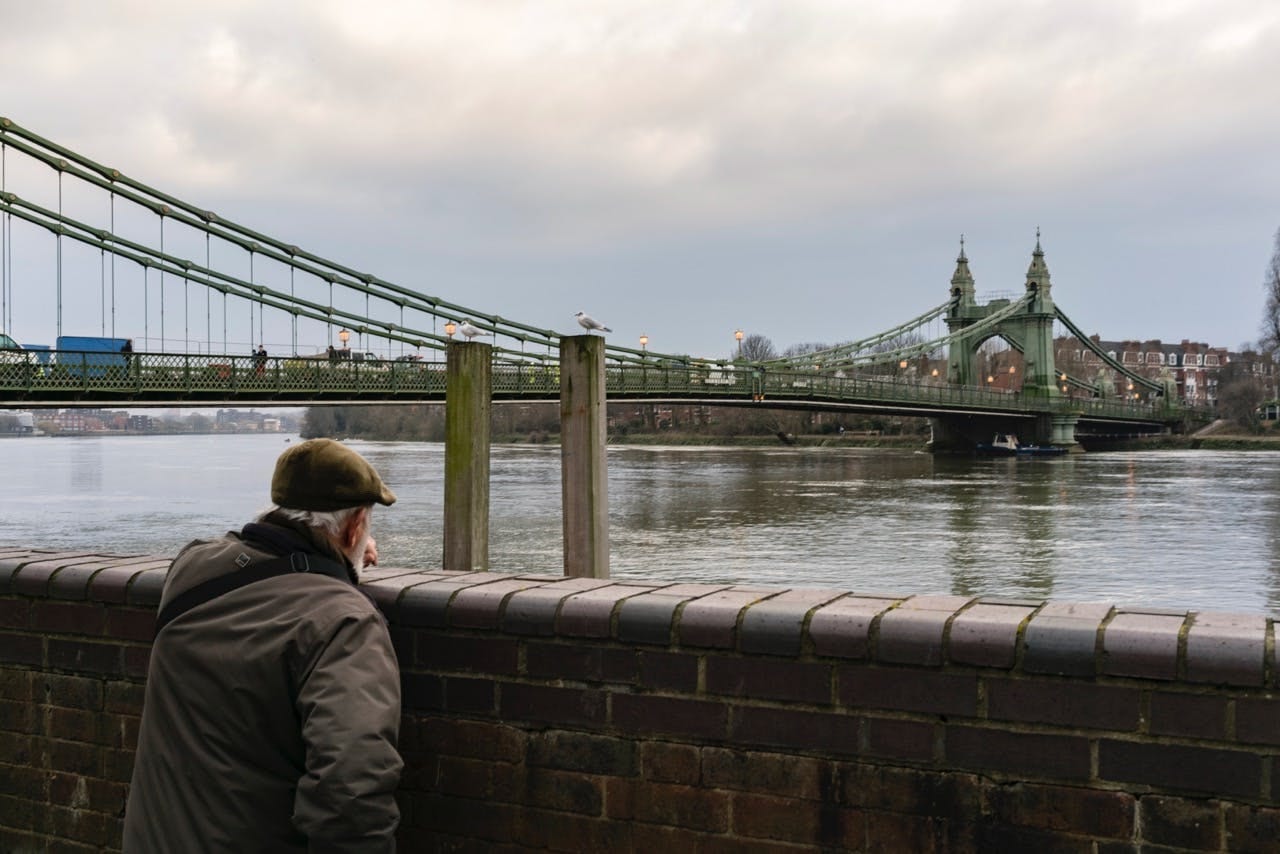 Elderly man in a flat cap gazing at Hammersmith Bridge across the Thames