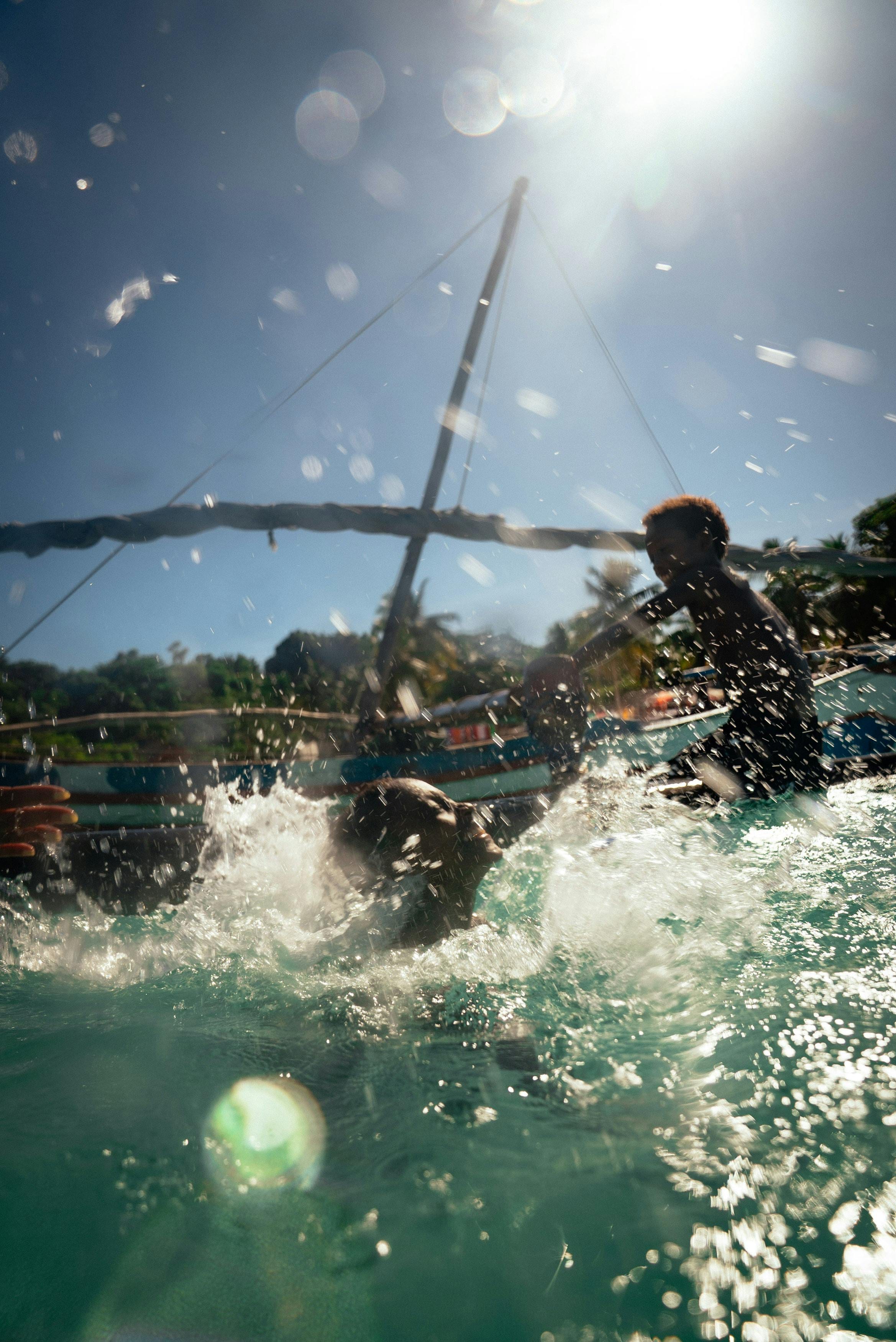 Children splashing beside a traditional sailing pirogue, sun flare over turquoise water
