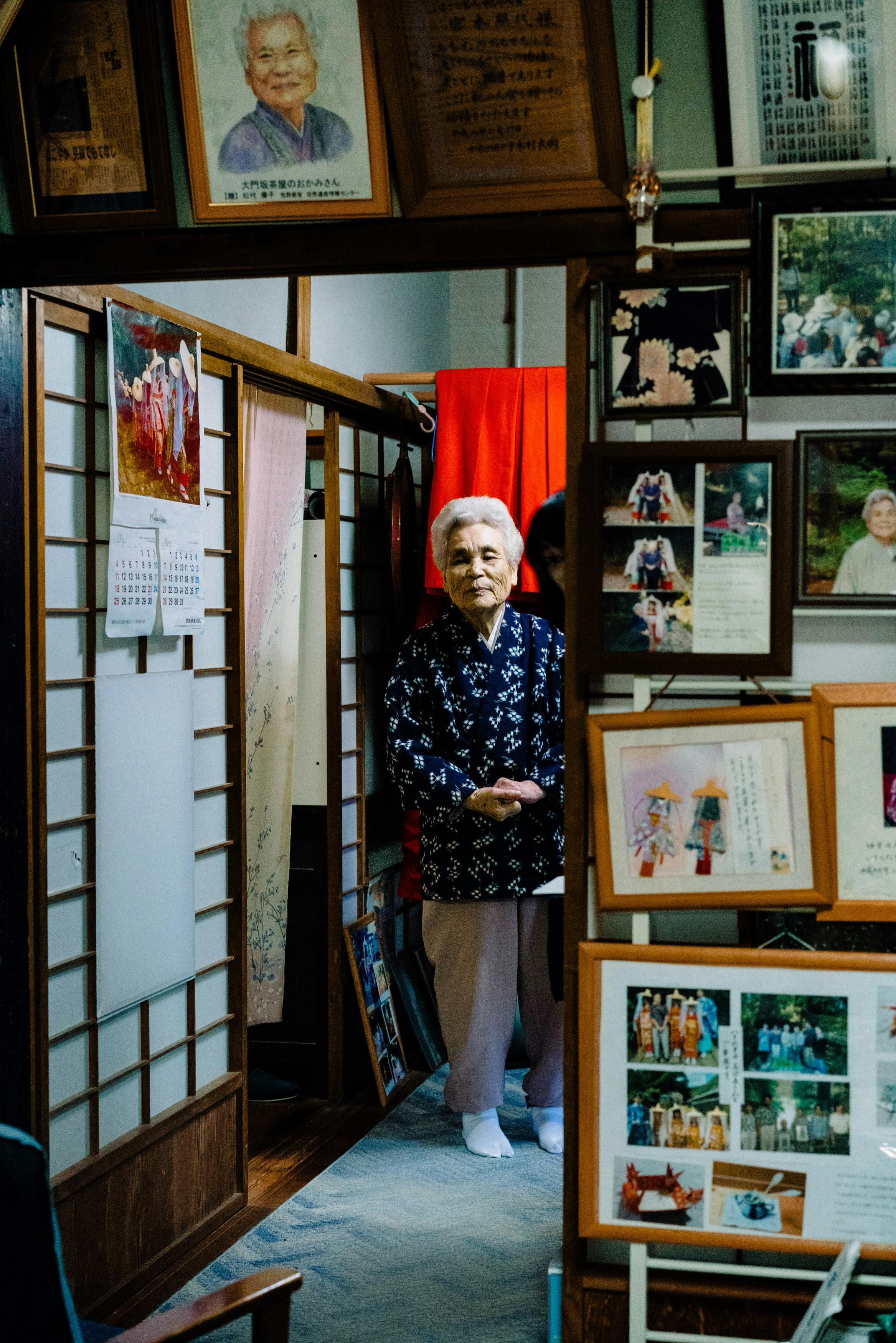 Elderly Japanese woman standing among framed photos in a traditional teahouse