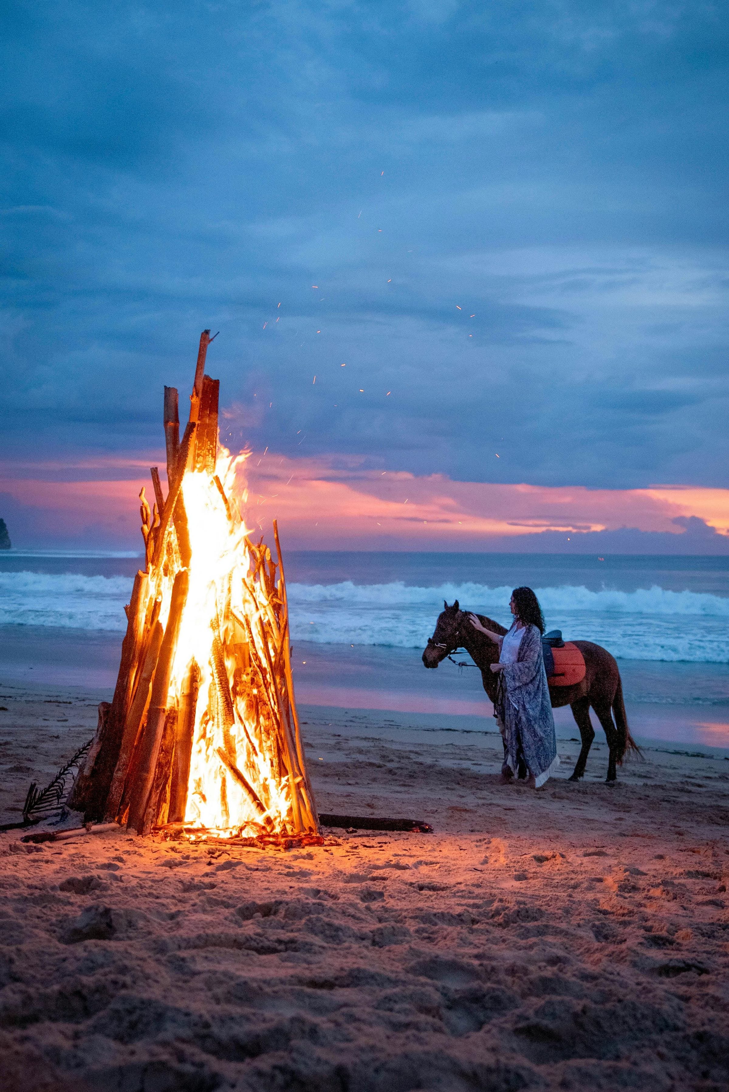 Woman with a saddled horse beside a towering driftwood bonfire on the beach at twilight