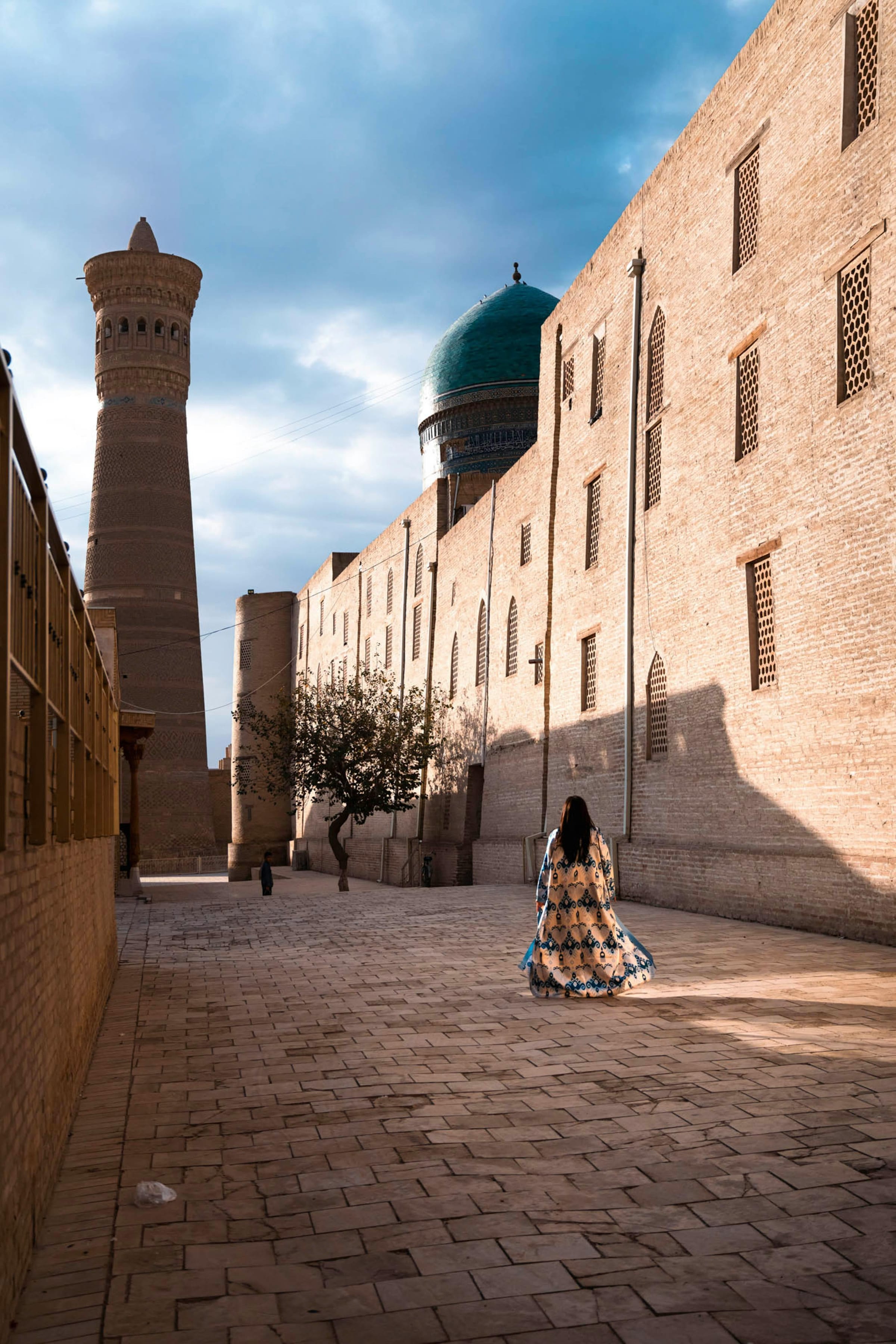 Woman in patterned robe walking an empty Bukhara lane toward the Kalyan minaret and turquoise dome